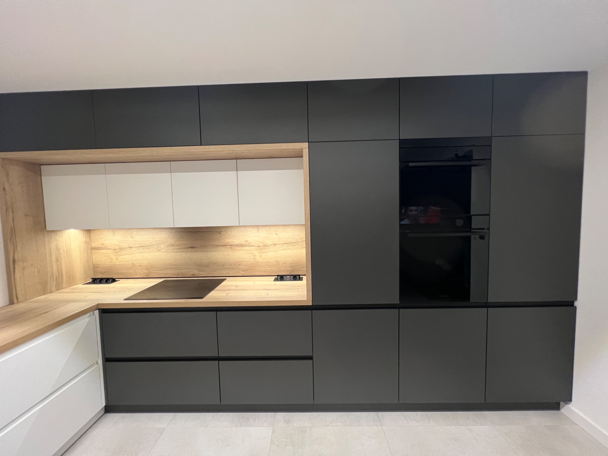 Nowoczesna kuchnia z czarnymi frontami i drewnianym blatem. Minimalistyczny design z wbudowanym piekarnikiem i płytą grzewczą. Jasne, neutralne tło.