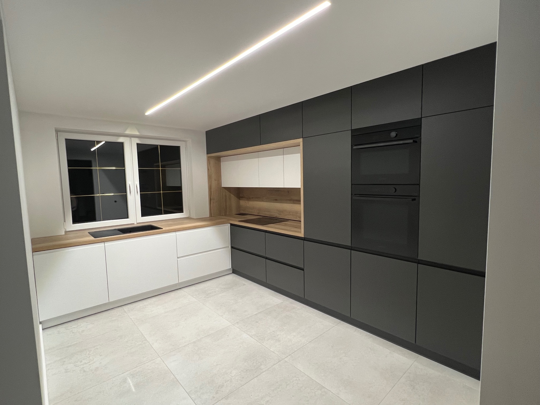 Nowoczesna kuchnia: grafitowe szafki z piekarnikiem w zabudowie, białe dolne szafki z drewnianym blatem, minimalistyczne oświetlenie LED i jasna podłoga.