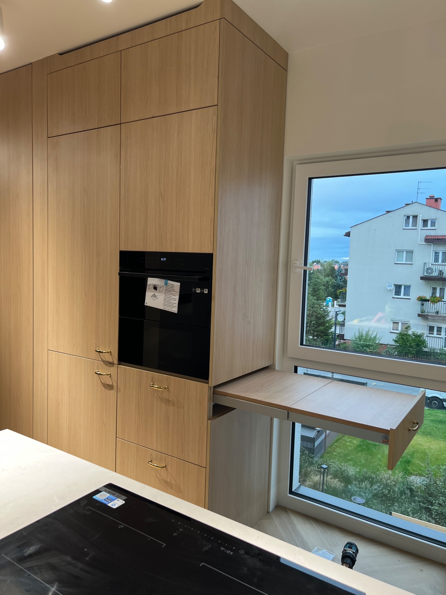 Jasna, drewniana zabudowa kuchenna z piekarnikiem i wysuwanym blatem przy oknie z widokiem na budynek mieszkalny. Minimalistyczne uchwyty, płyta indukcyjna na blacie.