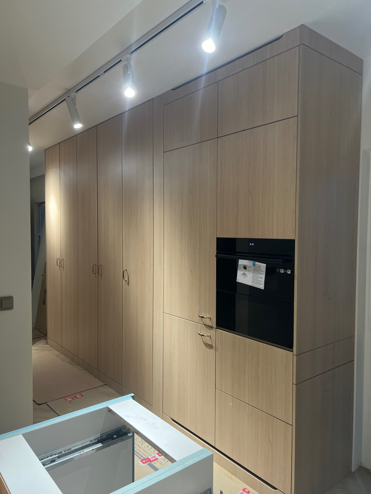 Jasna zabudowa kuchenna z piekarnikiem i szafkami, widok z dołu, fragment blatu roboczego w kadrze. Oświetlenie punktowe na suficie. Minimalistyczny design.