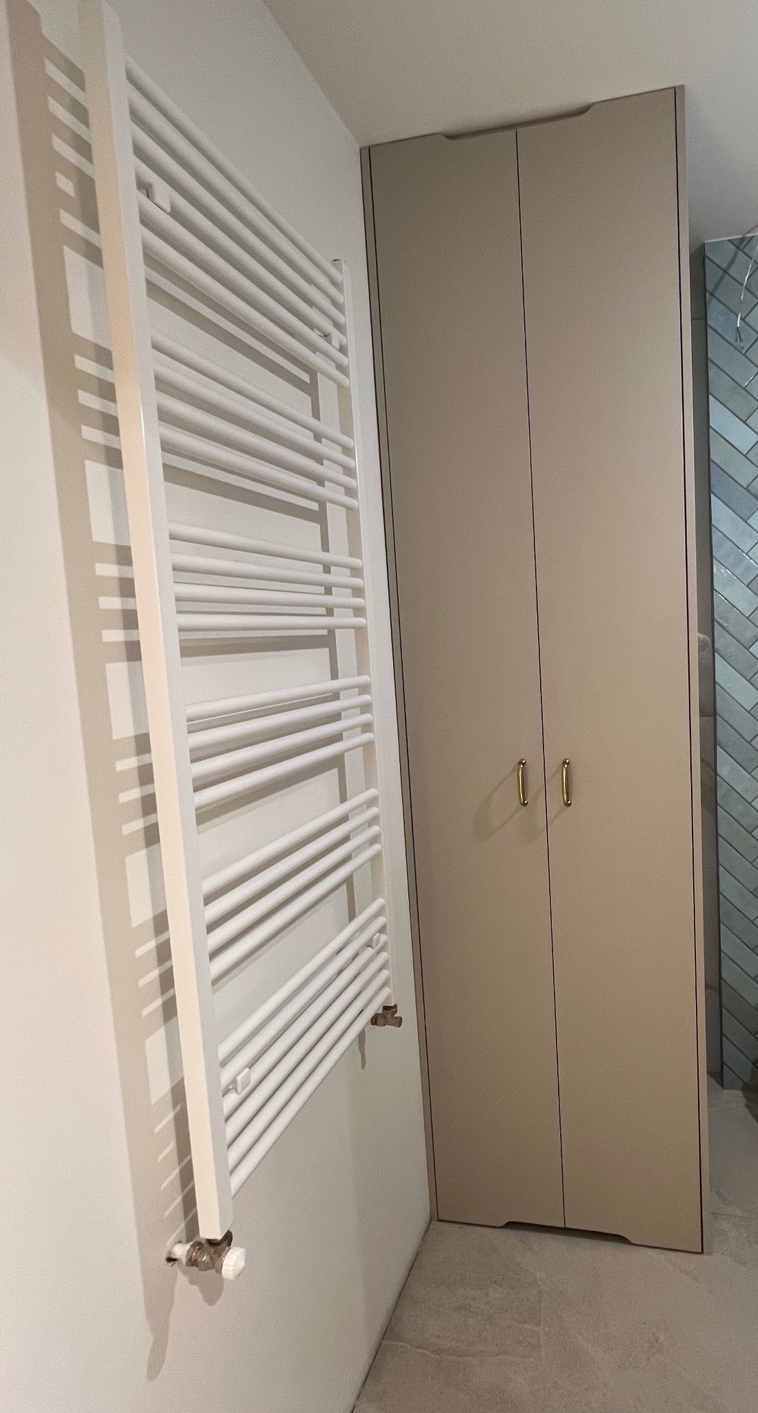 Beżowa szafa wnękowa z złotymi uchwytami, obok biały grzejnik drabinkowy w łazience z szarą podłogą i niebieskimi płytkami w jodełkę w tle.