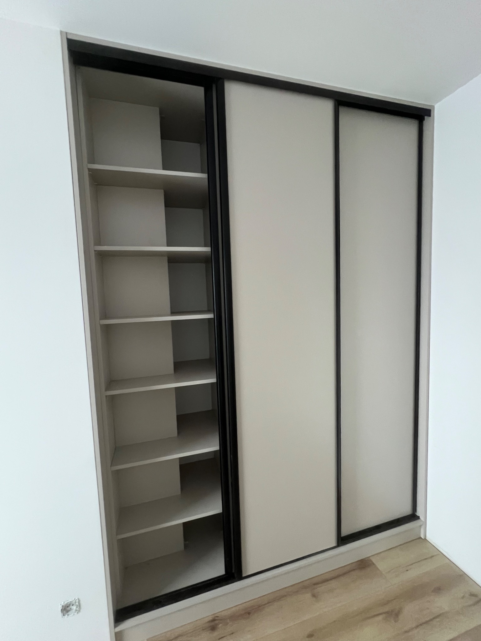 Jasna szafa wnękowa z przesuwnymi drzwiami i czarnymi ramami, z widocznymi półkami po jednej stronie. Minimalistyczny design, jasne wnętrze, drewniana podłoga.