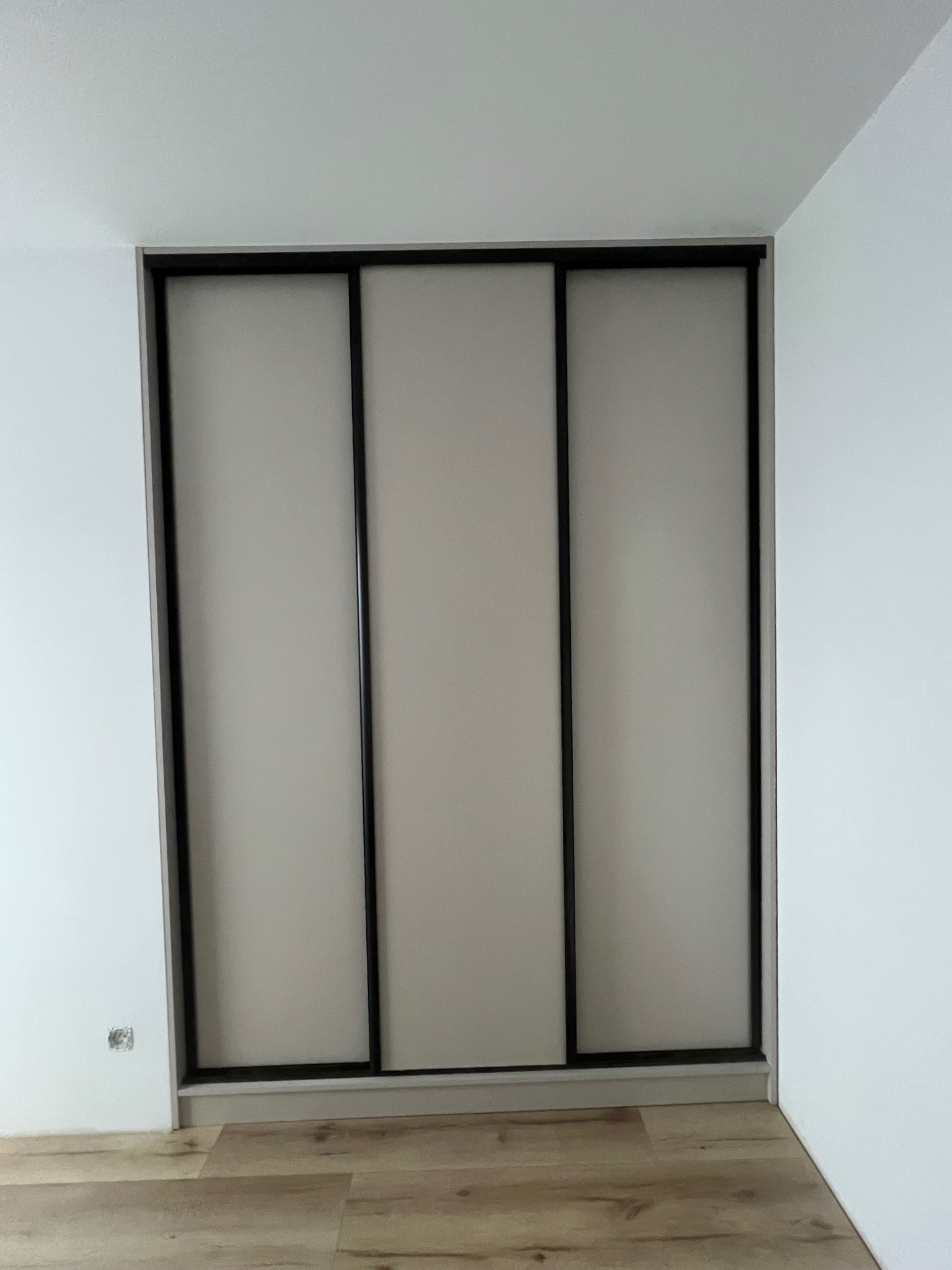 Minimalistyczna szafa wnękowa z trzema przesuwnymi drzwiami w odcieniach szarości, z czarnymi ramami, wkomponowana w białe ściany i jasną podłogę.