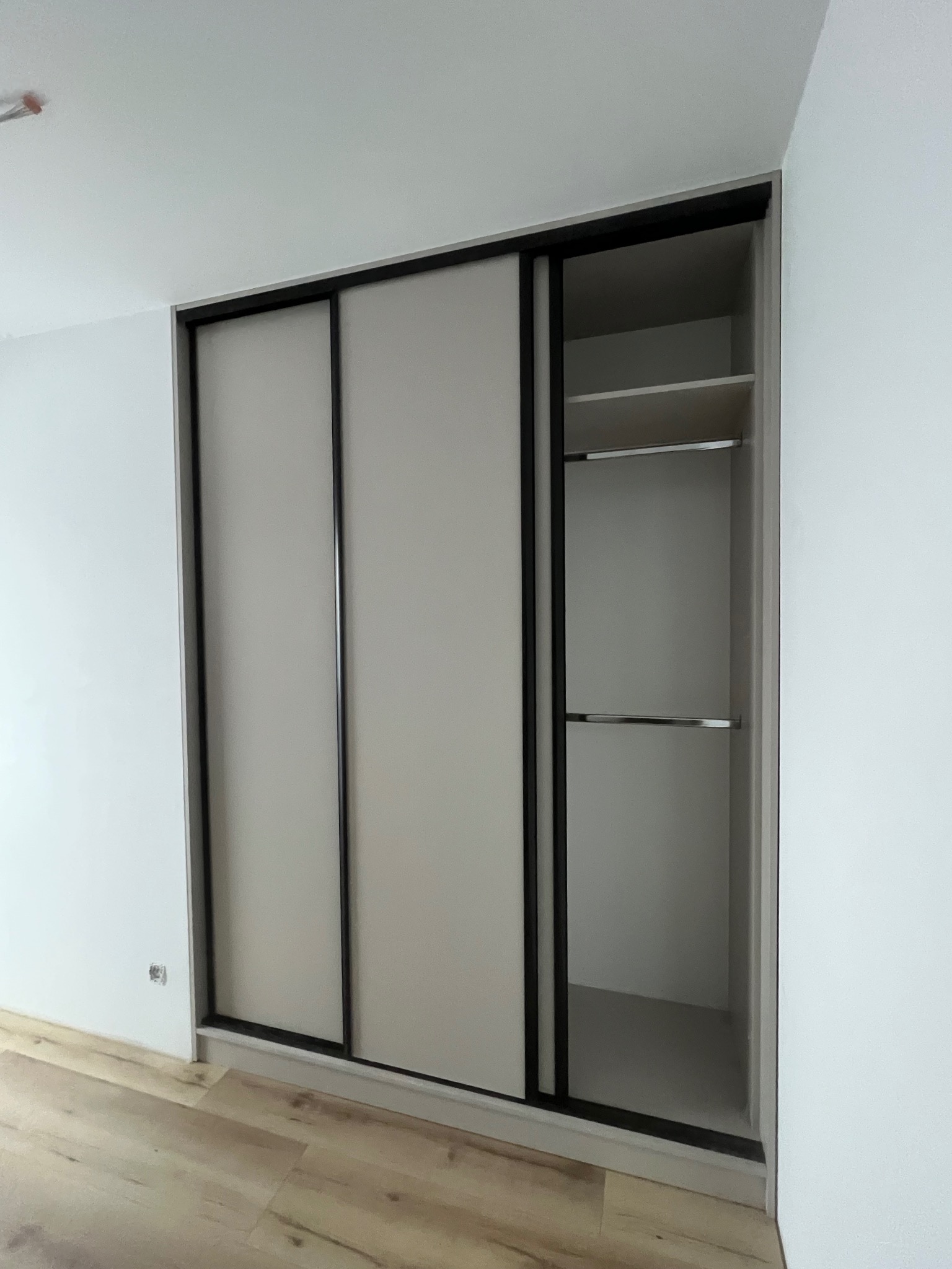 Jasna szafa wnękowa z przesuwnymi drzwiami i czarnymi obramowaniami, z otwartą częścią ukazującą półki z metalowymi drążkami, na tle białych ścian i drewnianej podłogi.