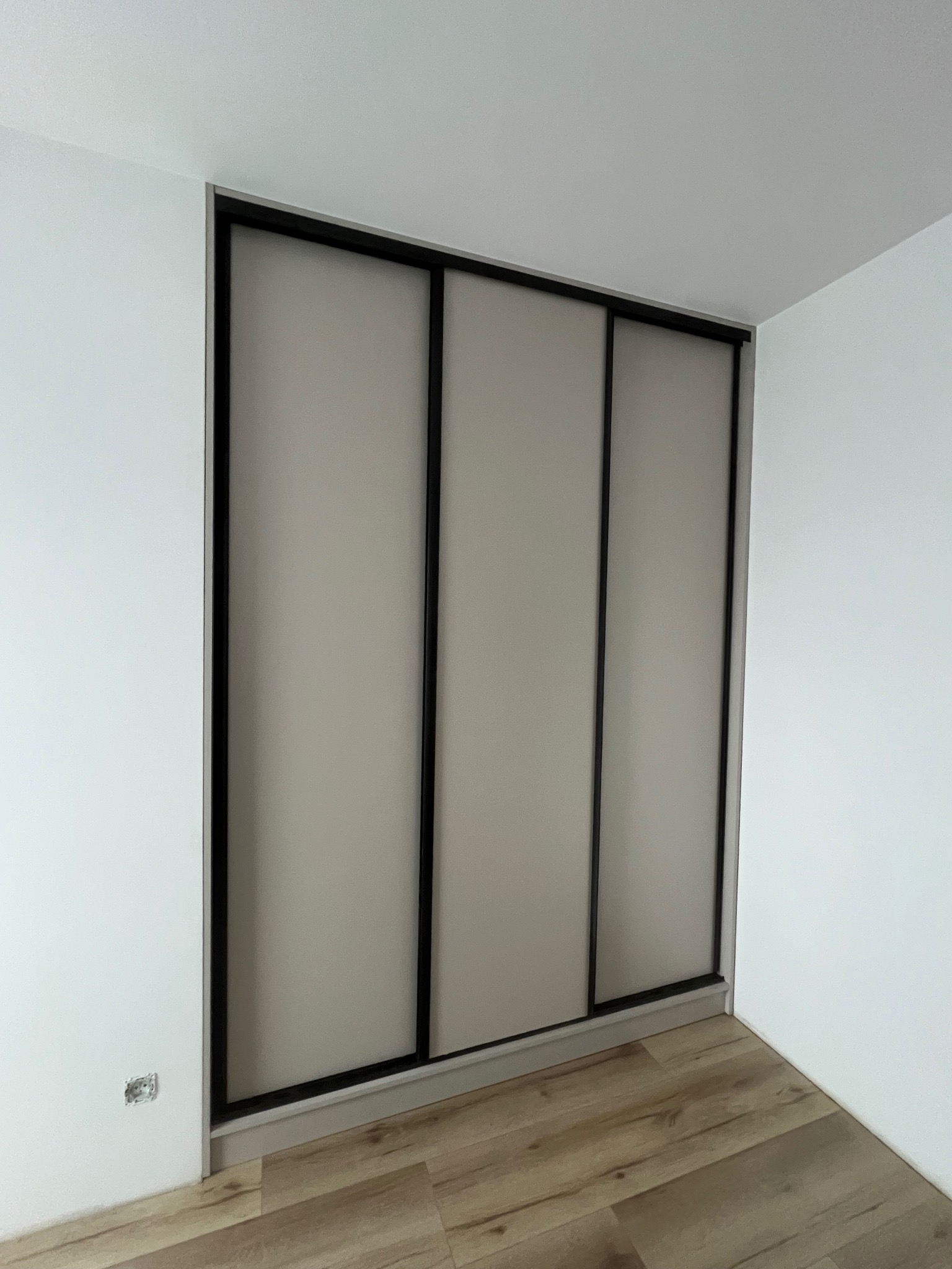 Wnętrze z szafą wnękową z przesuwnymi, beżowymi drzwiami z czarnymi ramami. Widoczna podłoga z paneli i białe ściany. Gniazdko elektryczne wystaje ze ściany.