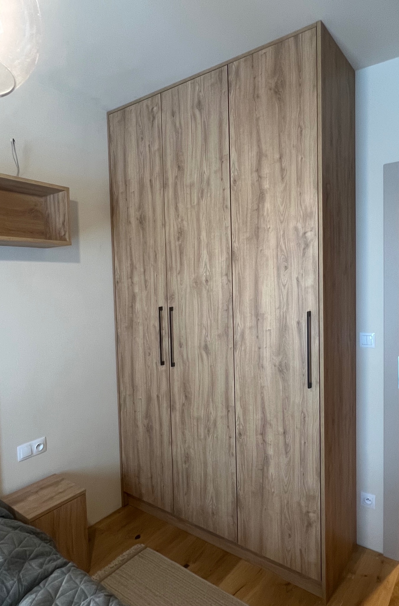 Trzydrzwiowa szafa na wymiar z drewnopodobnym fornirem, z długimi, pionowymi uchwytami, dopasowana do wnętrza sypialni z drewnianą podłogą i szafką nocną.