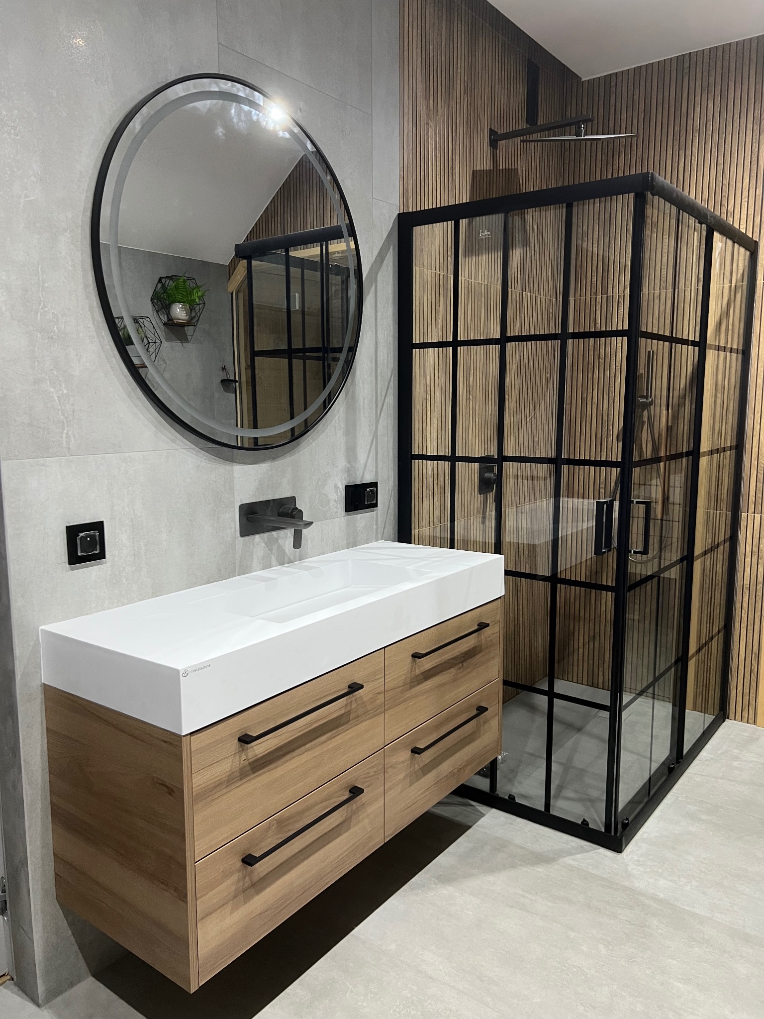 Nowoczesna łazienka w Kleczy Górnej: szafka pod umywalkę z drewnianymi frontami, okrągłe lustro i kabina prysznicowa z czarnymi szprosami na tle drewnianej ściany.