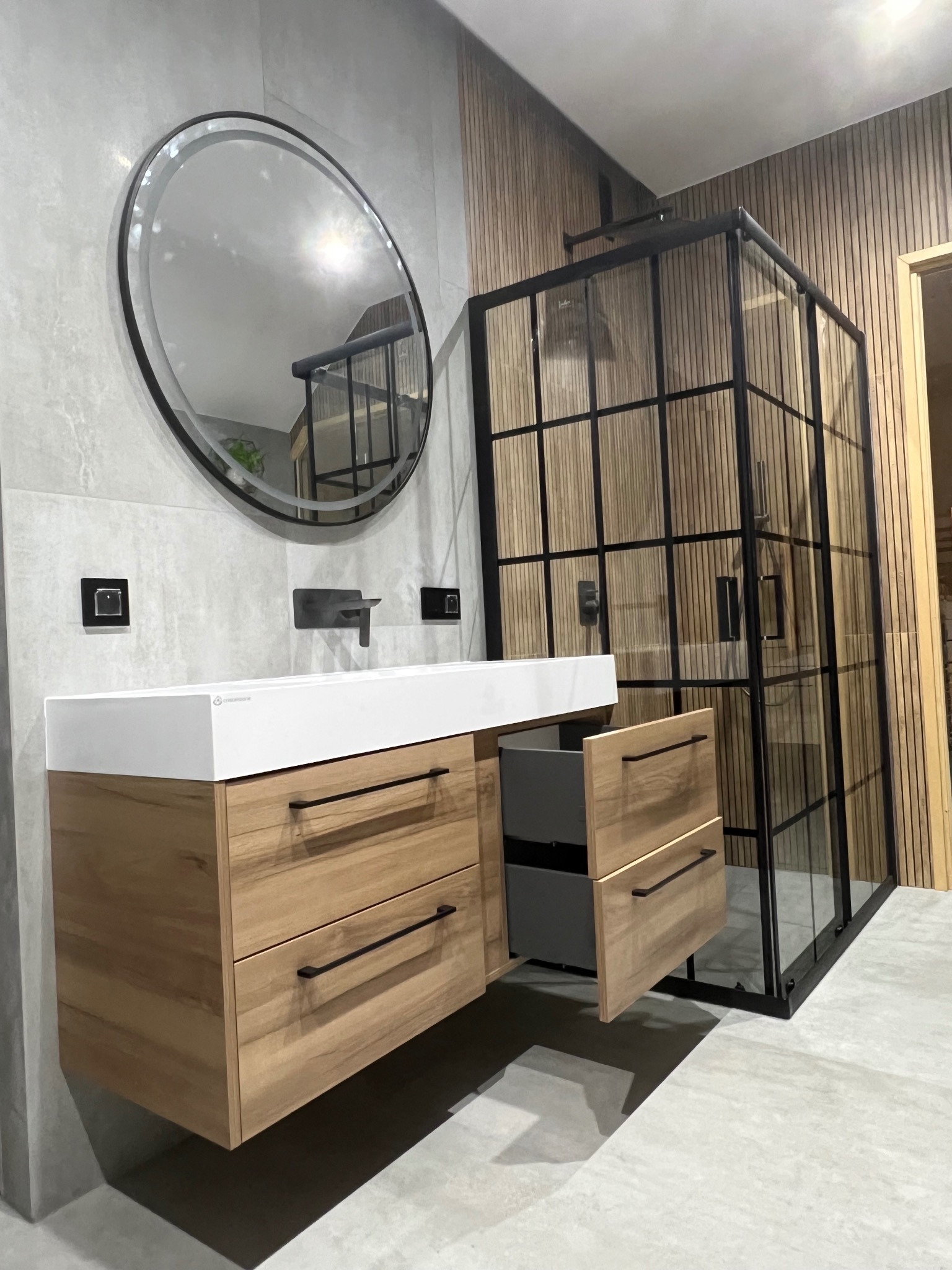 Nowoczesna łazienka z drewnianą szafką pod umywalkę z otwartą szufladą, okrągłym lustrem i kabiną prysznicową w stylu loft z czarnymi szprosami.