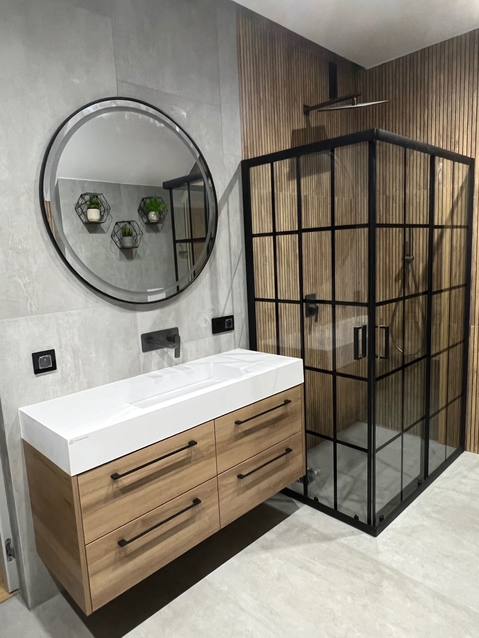 Nowoczesna łazienka z drewnianą zabudową umywalki z białym blatem, okrągłym lustrem i kabiną prysznicową w stylu loft na tle szarych i drewnianych płytek.