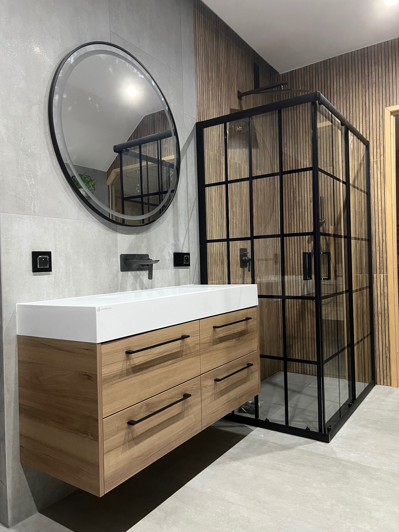 Nowoczesna łazienka: drewniana szafka pod umywalką, okrągłe lustro i kabina prysznicowa z czarnymi ramami na tle drewnianej ściany.