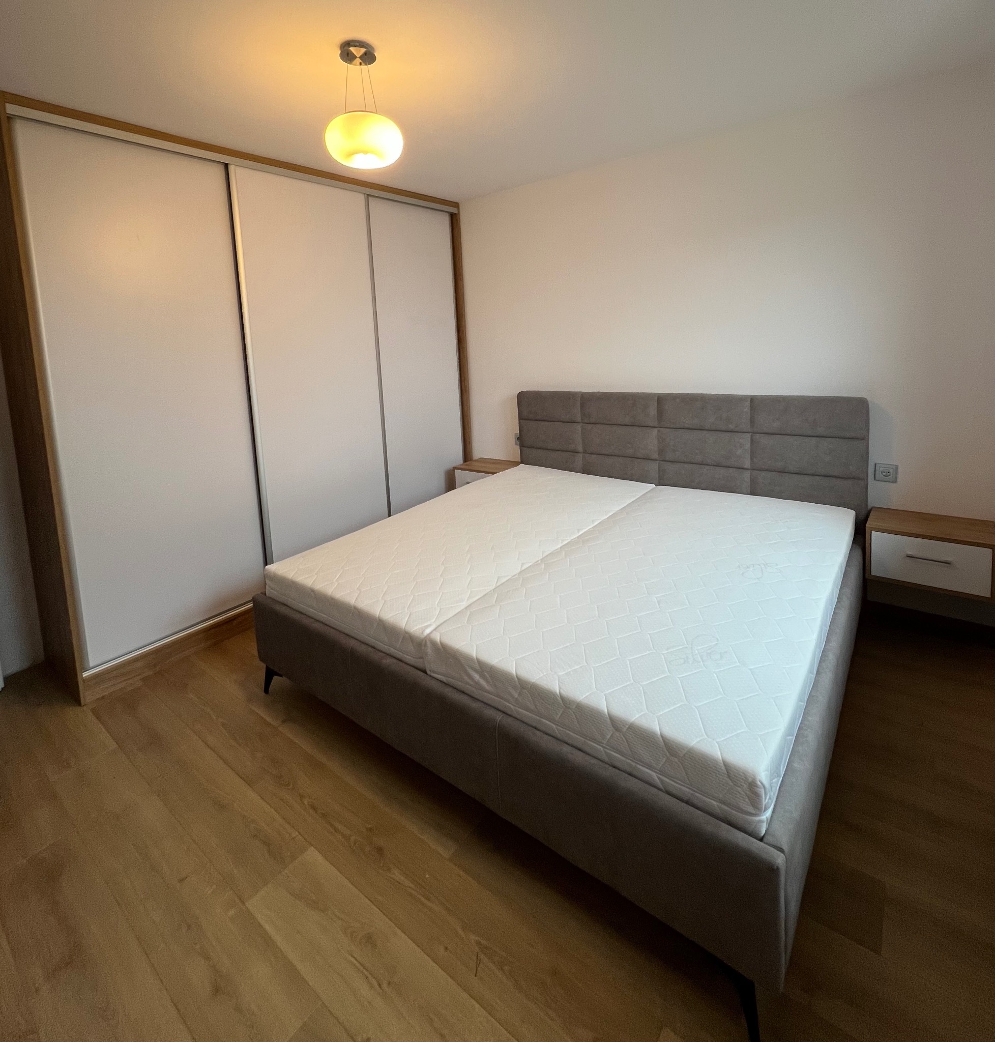 Nowoczesna szafa przesuwna z białymi frontami i drewnianą ramą, obok łóżko z szarym zagłówkiem i szafką nocną. Ciepłe oświetlenie sufitowe i drewniana podłoga.