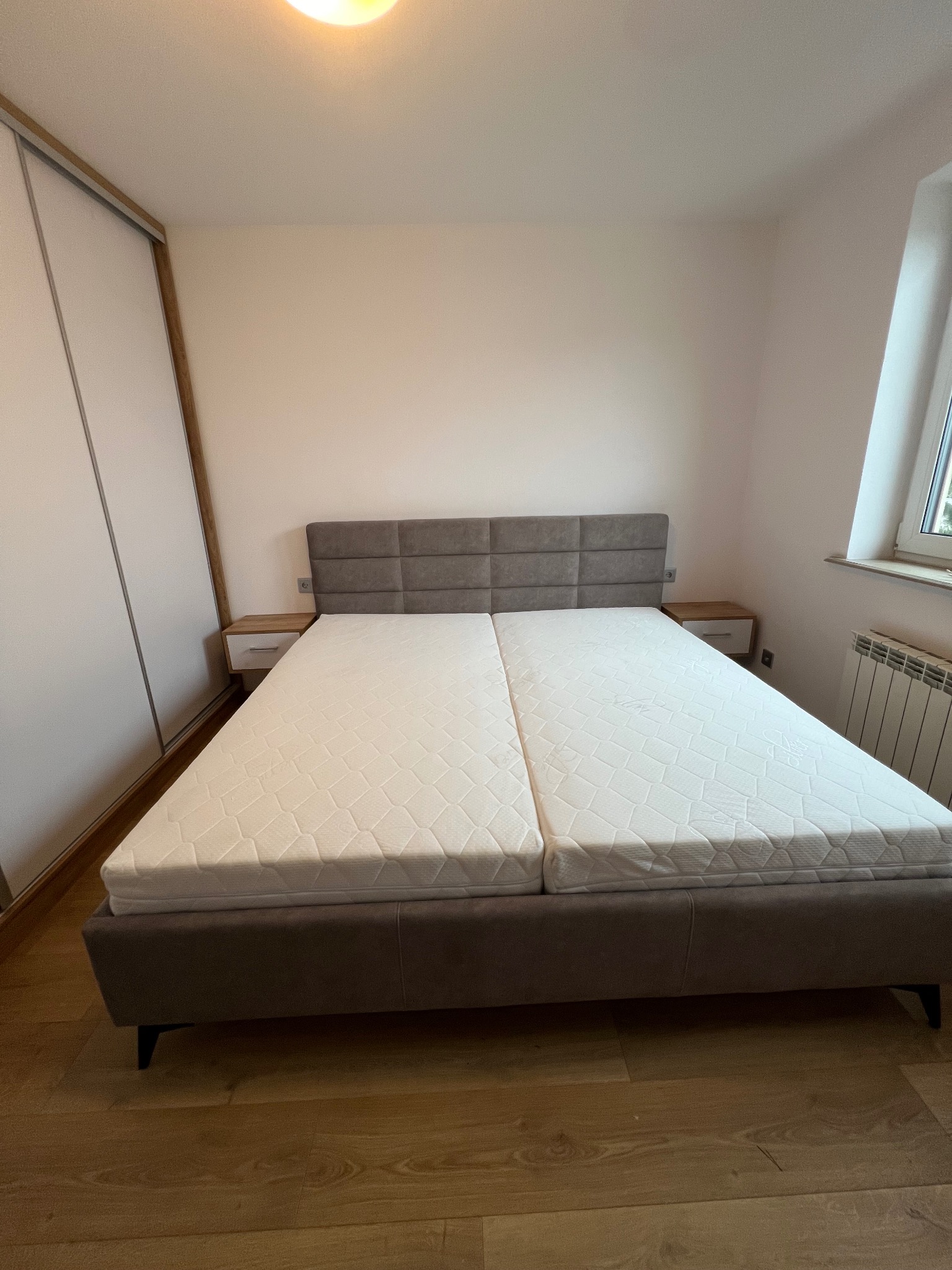 Nowoczesne łóżko z szarym tapicerowanym zagłówkiem i dwiema oddzielnymi nakładkami na materac, z szafkami nocnymi po bokach, w jasnym pokoju z drewnianą podłogą.