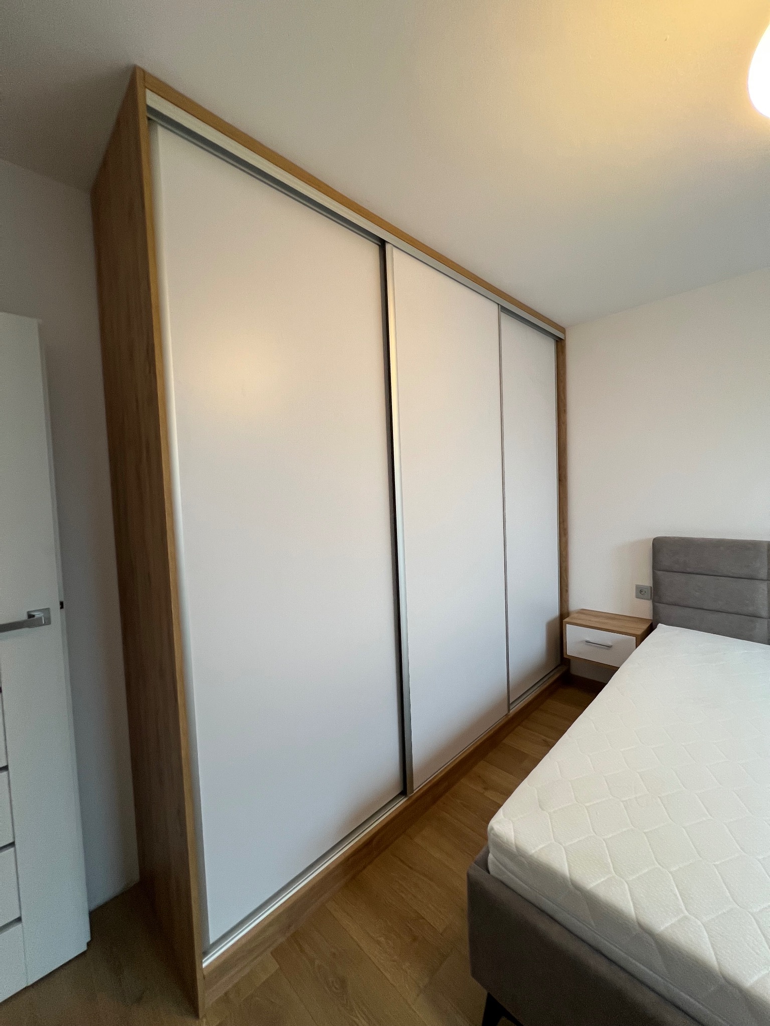 Szafa wnękowa z przesuwnymi, białymi drzwiami i drewnianym obramowaniem, widoczna w sypialni obok łóżka i szafki nocnej. Ujęcie z dołu, podkreślające wysokość mebla.