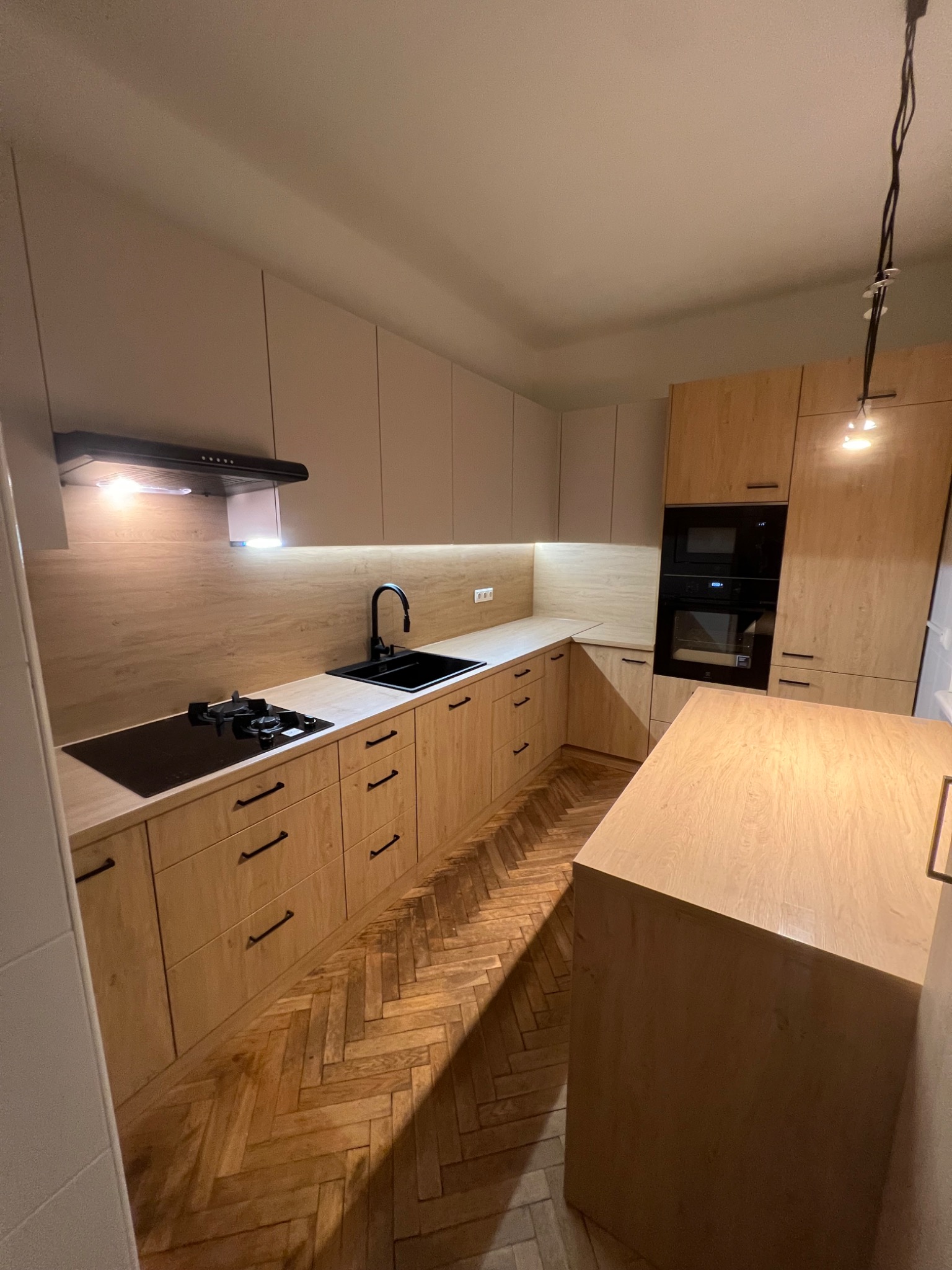 Jasna kuchnia z drewnianymi frontami i czarnymi detalami: zlew, bateria, płyta grzewcza. Parkiet w jodełkę. Oświetlenie podszafkowe i wisząca lampa.