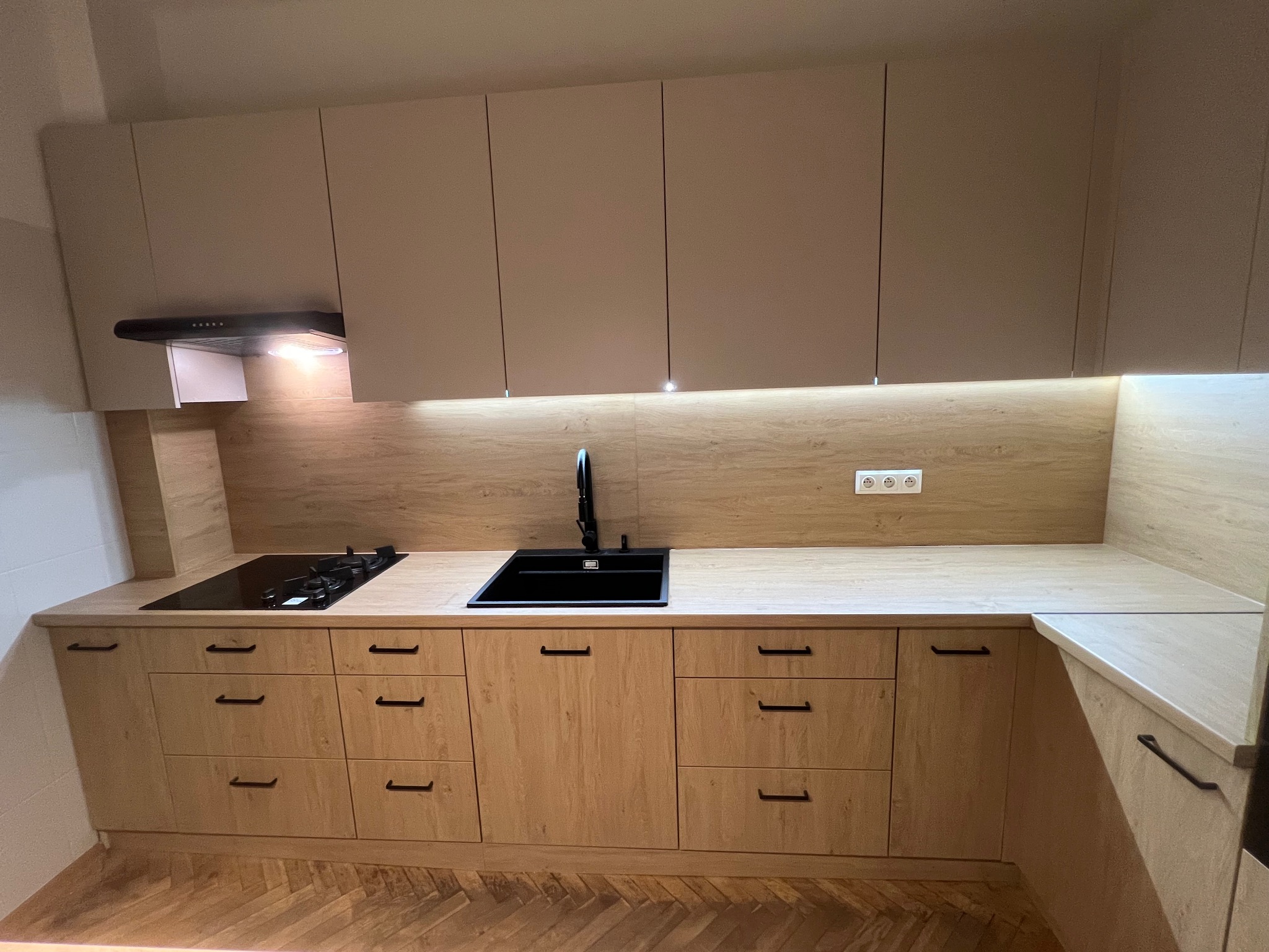 Jasna kuchnia z drewnianymi frontami i czarnymi uchwytami. Uwagę przykuwa czarny zlew i płyta grzewcza wkomponowana w blat. Podświetlenie LED dodaje nowoczesności.