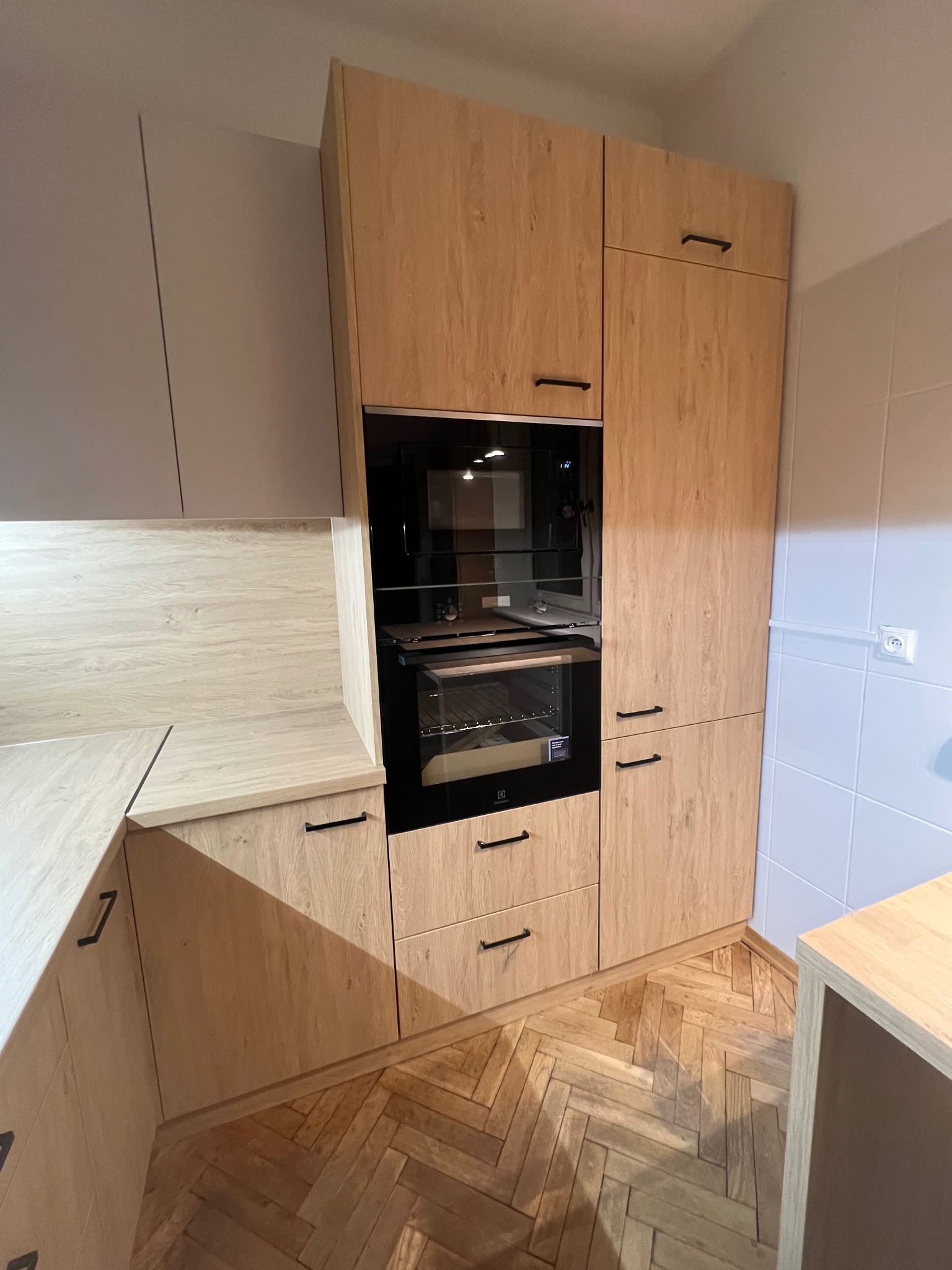 Jasna, drewnopodobna zabudowa kuchenna z piekarnikiem i czarnymi uchwytami, na tle parkietu ułożonego w jodełkę. Minimalistyczny design, nowoczesne wykończenie.