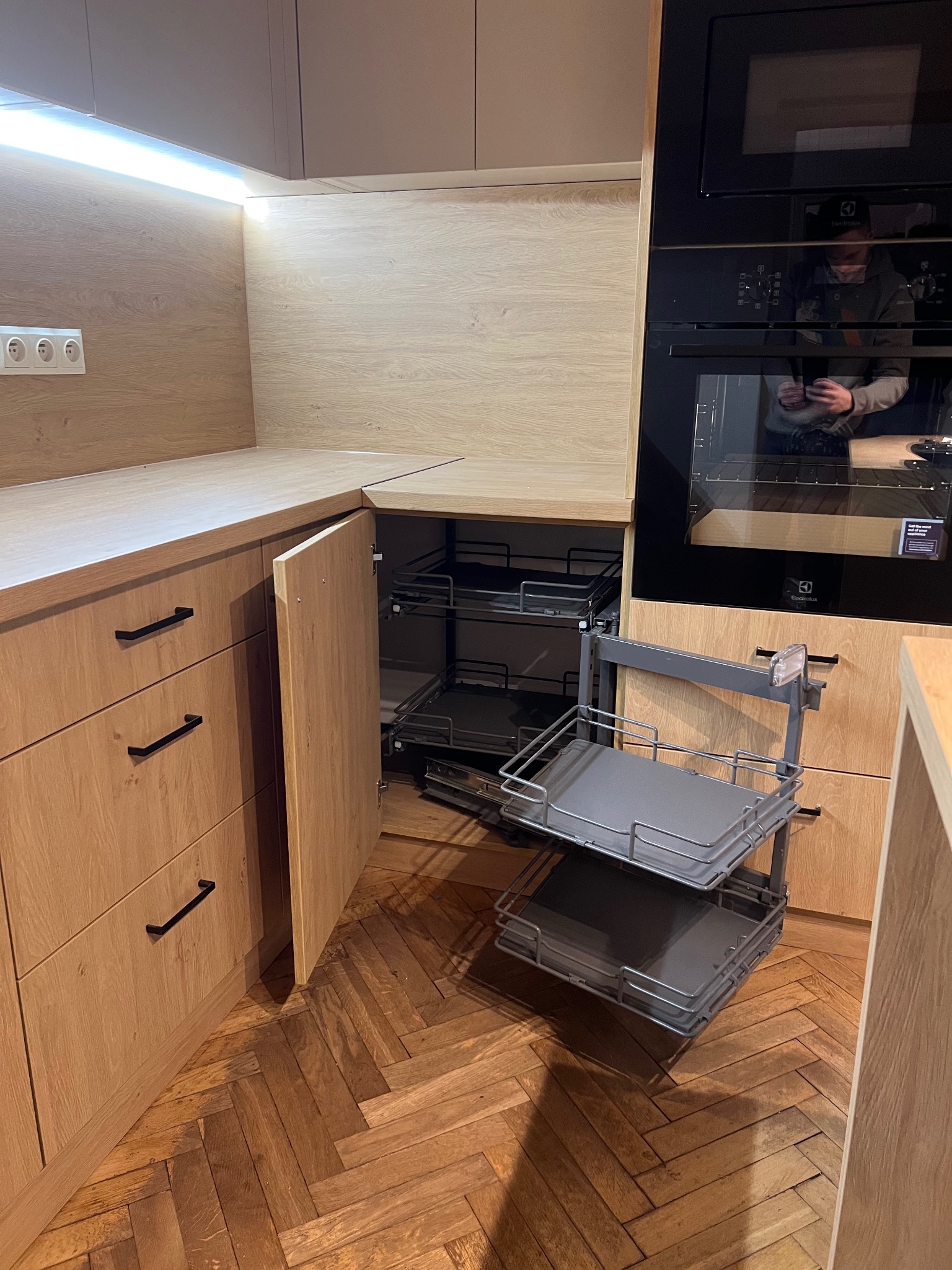 Jasna kuchnia z drewnianymi frontami, wysuwany kosz cargo. Widoczny piekarnik Electrolux i parkiet w jodełkę. Oświetlenie LED podszafkowe.