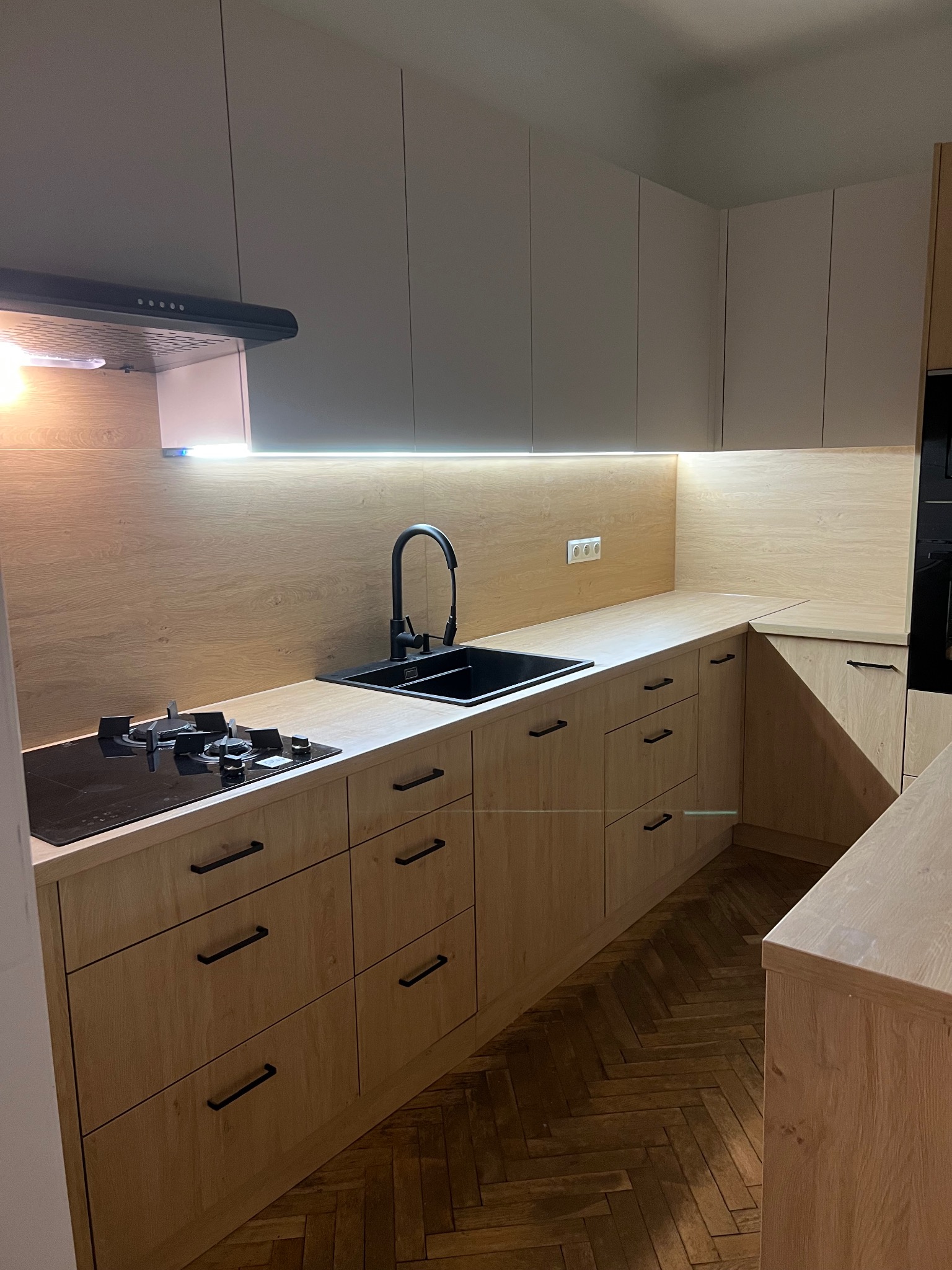 Jasna kuchnia z drewnianymi frontami i czarnymi uchwytami, oświetlona ledami. Widoczny zlew, płyta grzewcza i parkiet w jodełkę. Minimalistyczny design.