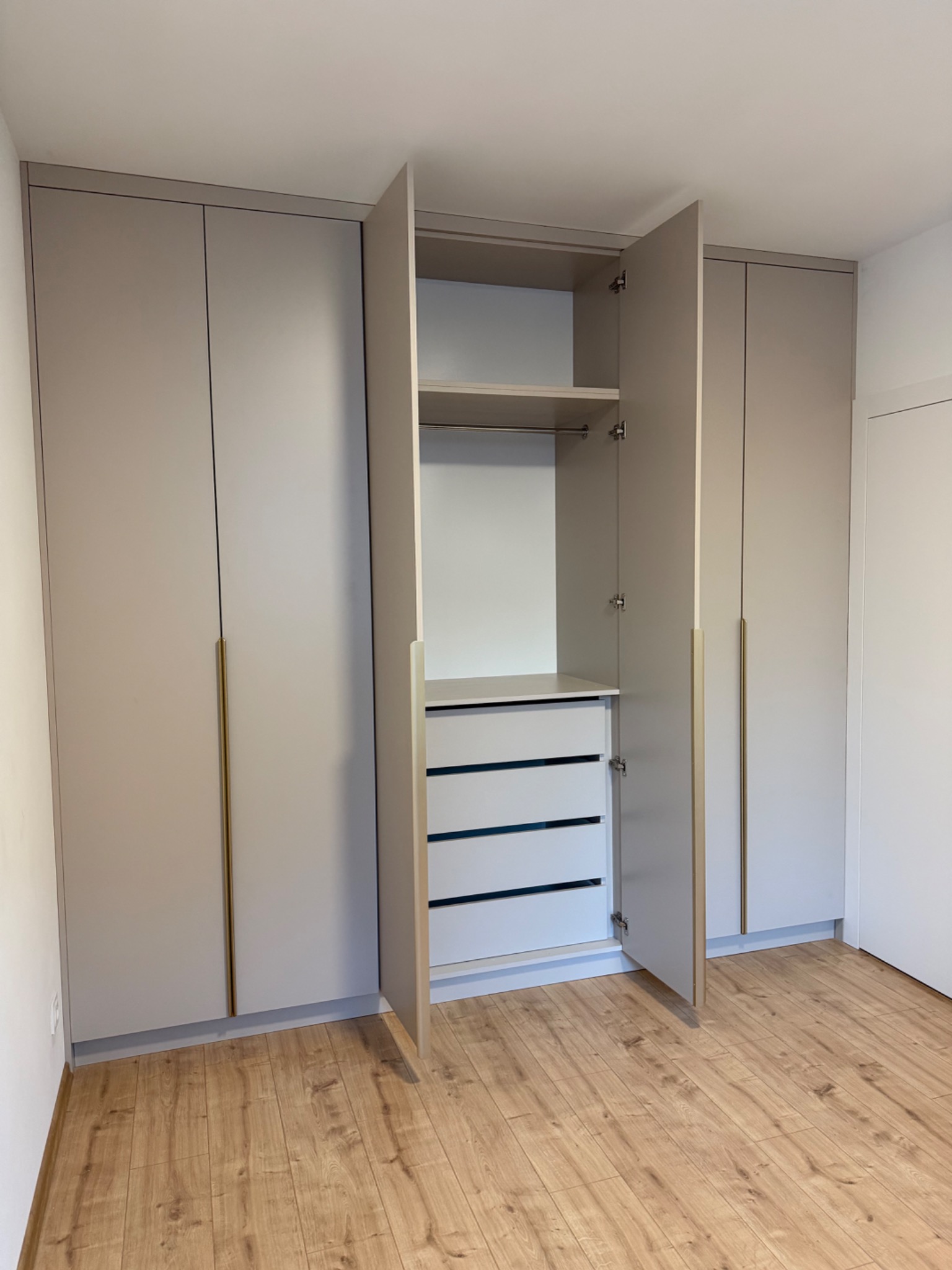 Jasnoszara szafa wnękowa z złotymi uchwytami, częściowo otwarta, ukazująca półki i szuflady. Na podłodze panele imitujące drewno. Minimalistyczny design.