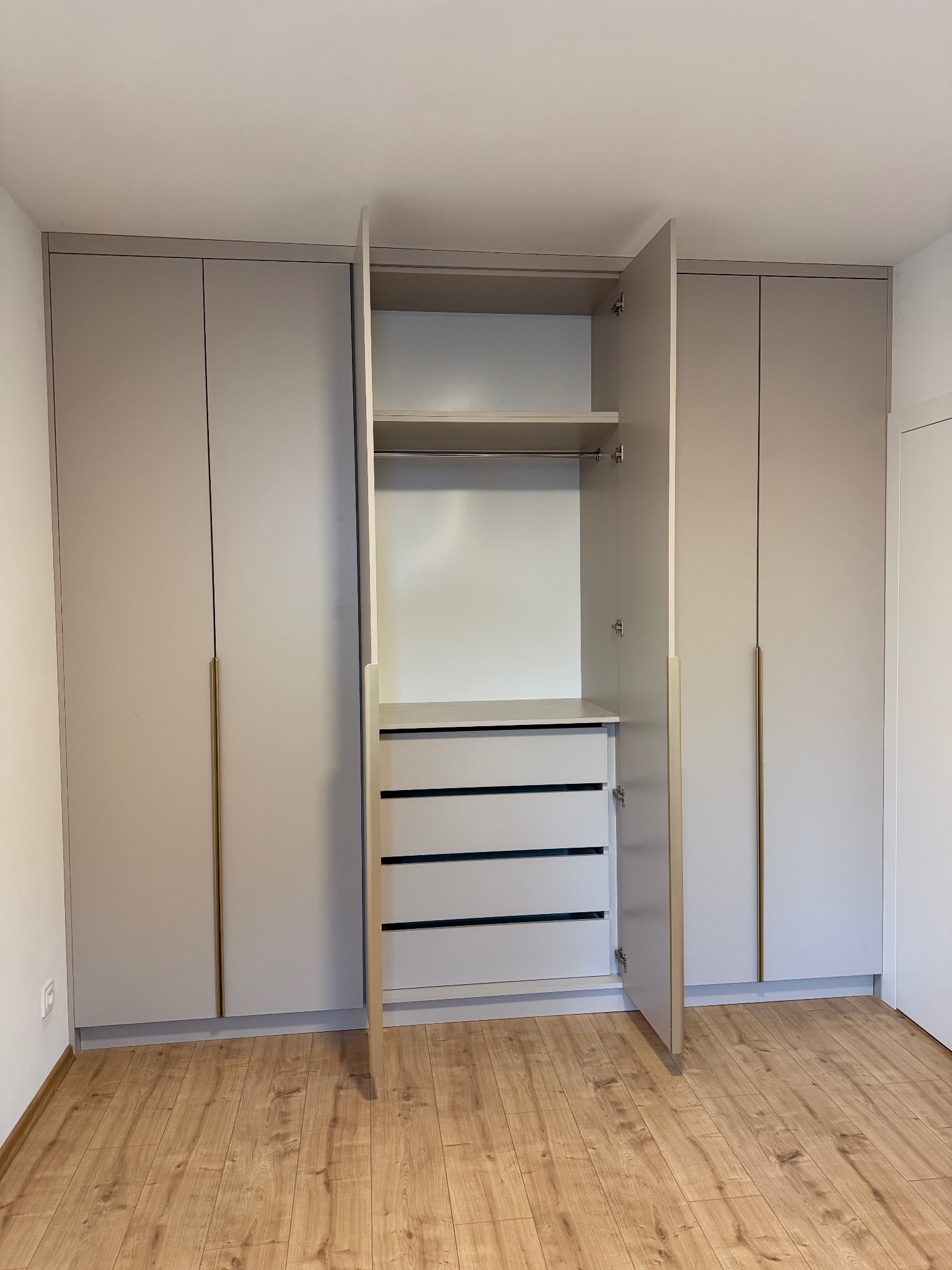 Jasnoszara szafa wnękowa z długimi, złotymi uchwytami. Jedne drzwi otwarte, ukazują półki i szuflady. Parkiet na podłodze. Minimalistyczny design.