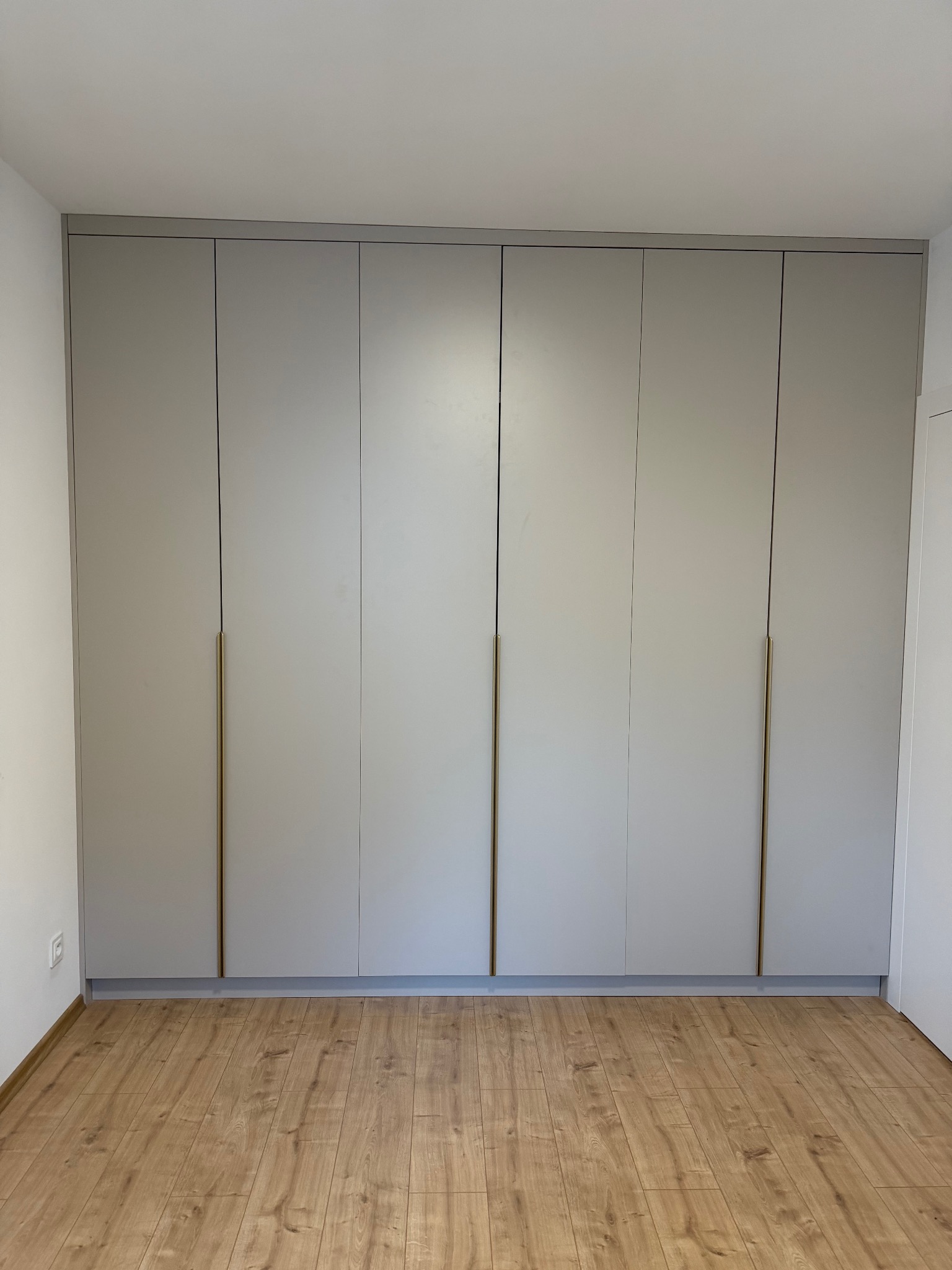 Jasnoszara szafa wnękowa z długimi, złotymi uchwytami, dopasowana do podłogi z paneli. Minimalistyczny design, widok na całą ścianę.