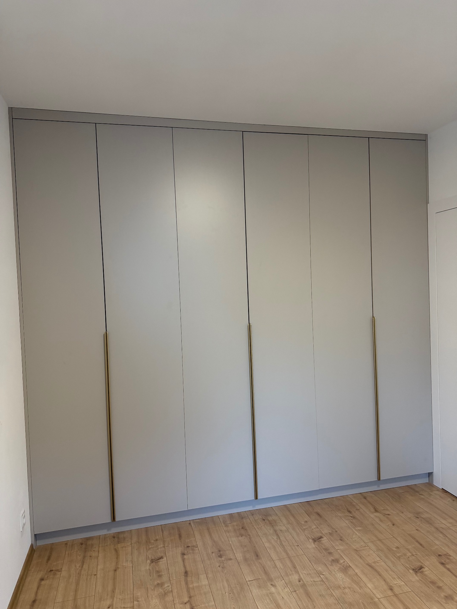 Jasnoszara szafa wnękowa z sześcioma drzwiami i długimi, złotymi uchwytami, dopasowana do sufitu, na tle parkietu. Minimalistyczny design.