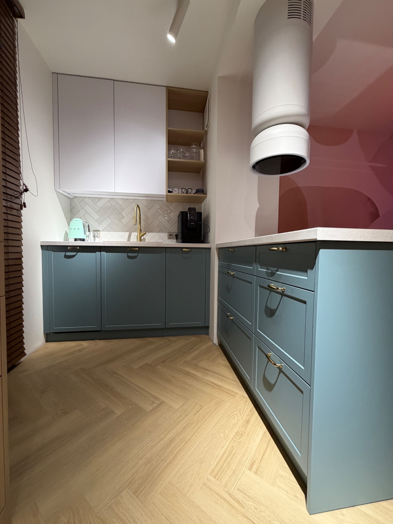 Niebieska kuchnia na wymiar z drewnianą podłogą w jodełkę. Białe szafki wiszące i złota armatura. Nowoczesny okap cylindryczny. Minimalistyczny design.