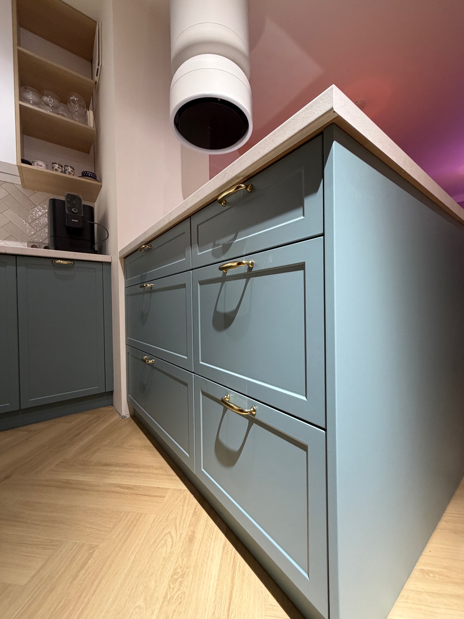 Niebieska zabudowa kuchenna z czterema szufladami i złotymi uchwytami, blat z jasnego kamienia, parkiet w jodełkę, nowoczesny okap cylindryczny.