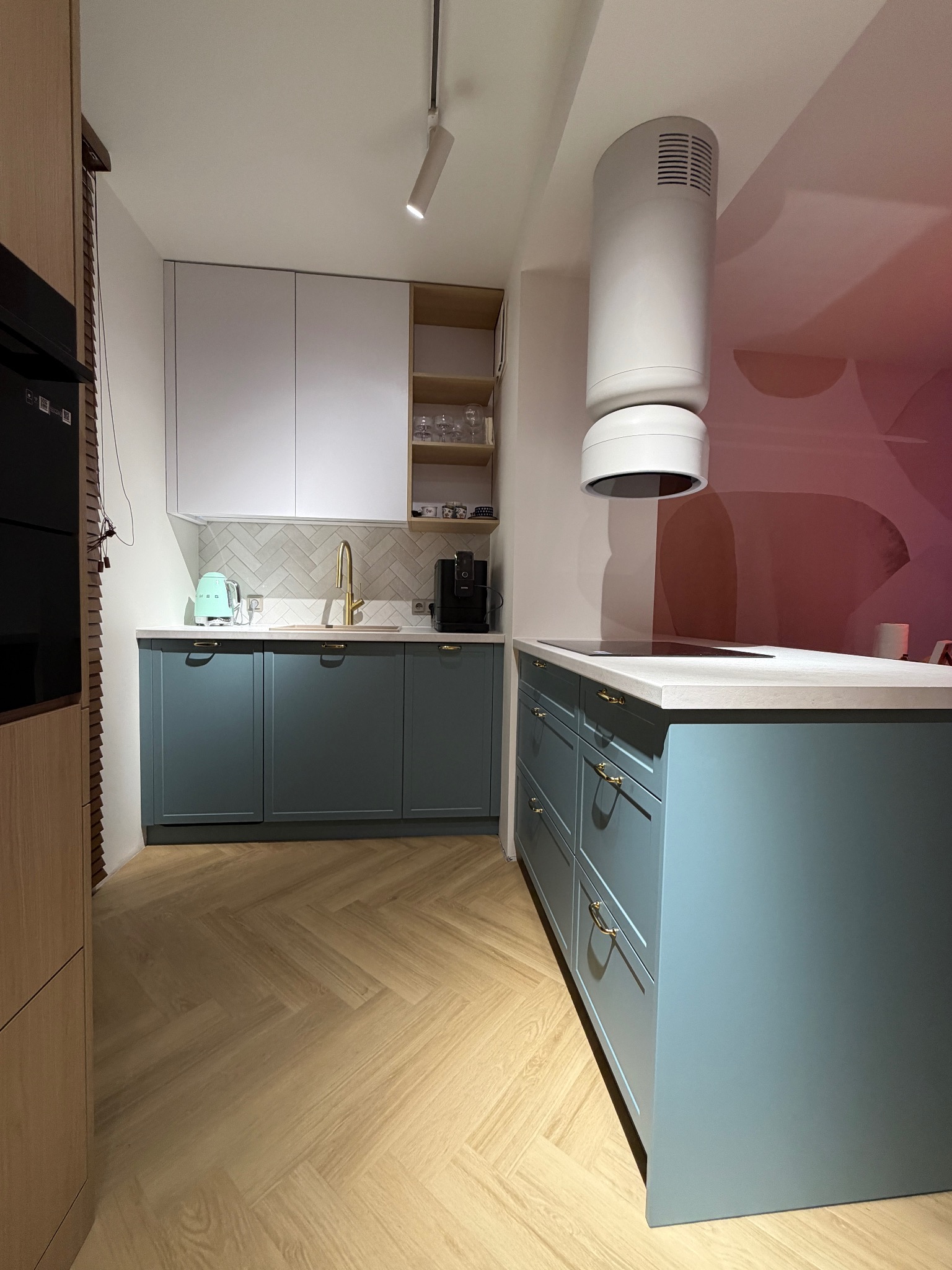 Nowoczesna kuchnia z niebieskimi szafkami, złotą armaturą i drewnianą podłogą w jodełkę. Białe szafki wiszące i designerski okap dodają elegancji.