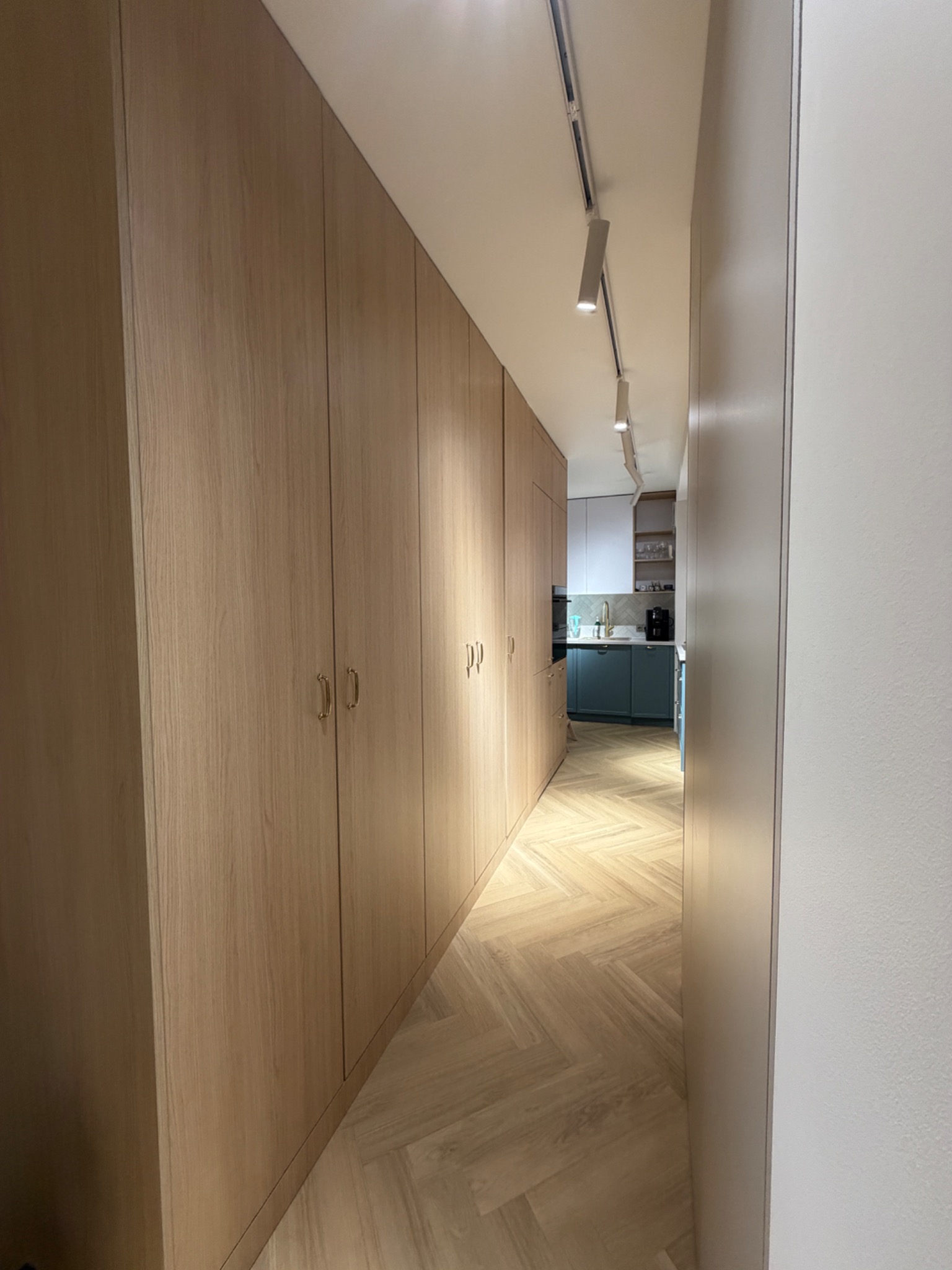 Długi korytarz z zabudową szaf w odcieniach drewna, parkiet w jodełkę i widok na kuchnię z niebieskimi szafkami. Oświetlenie szynowe sufitowe.