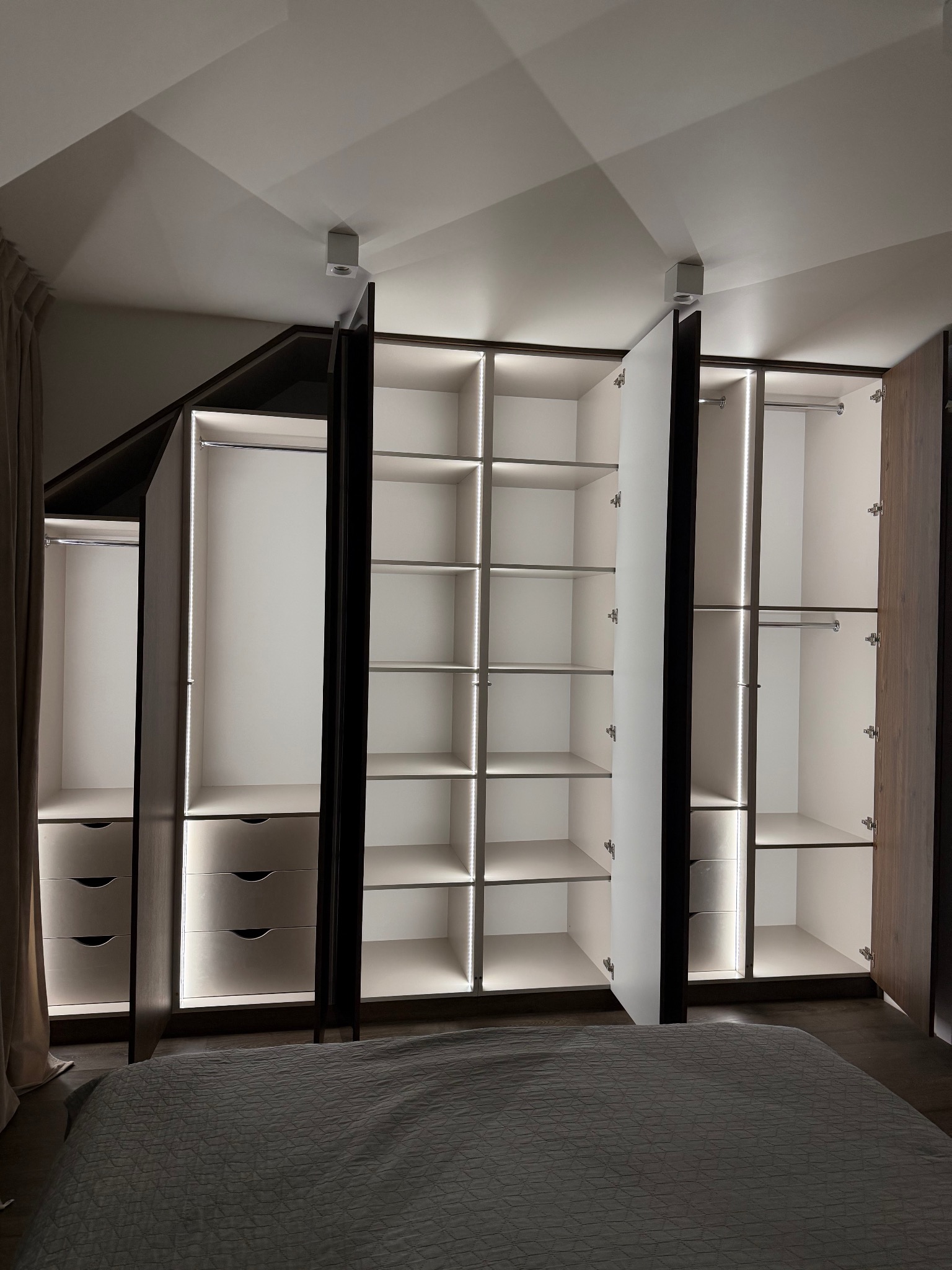 Nowoczesna szafa wnękowa z oświetleniem LED, widok z perspektywy łóżka. Częściowo otwarte drzwi ukazują półki i szuflady. Ciemne obramowanie kontrastuje z jasnym wnętrzem.