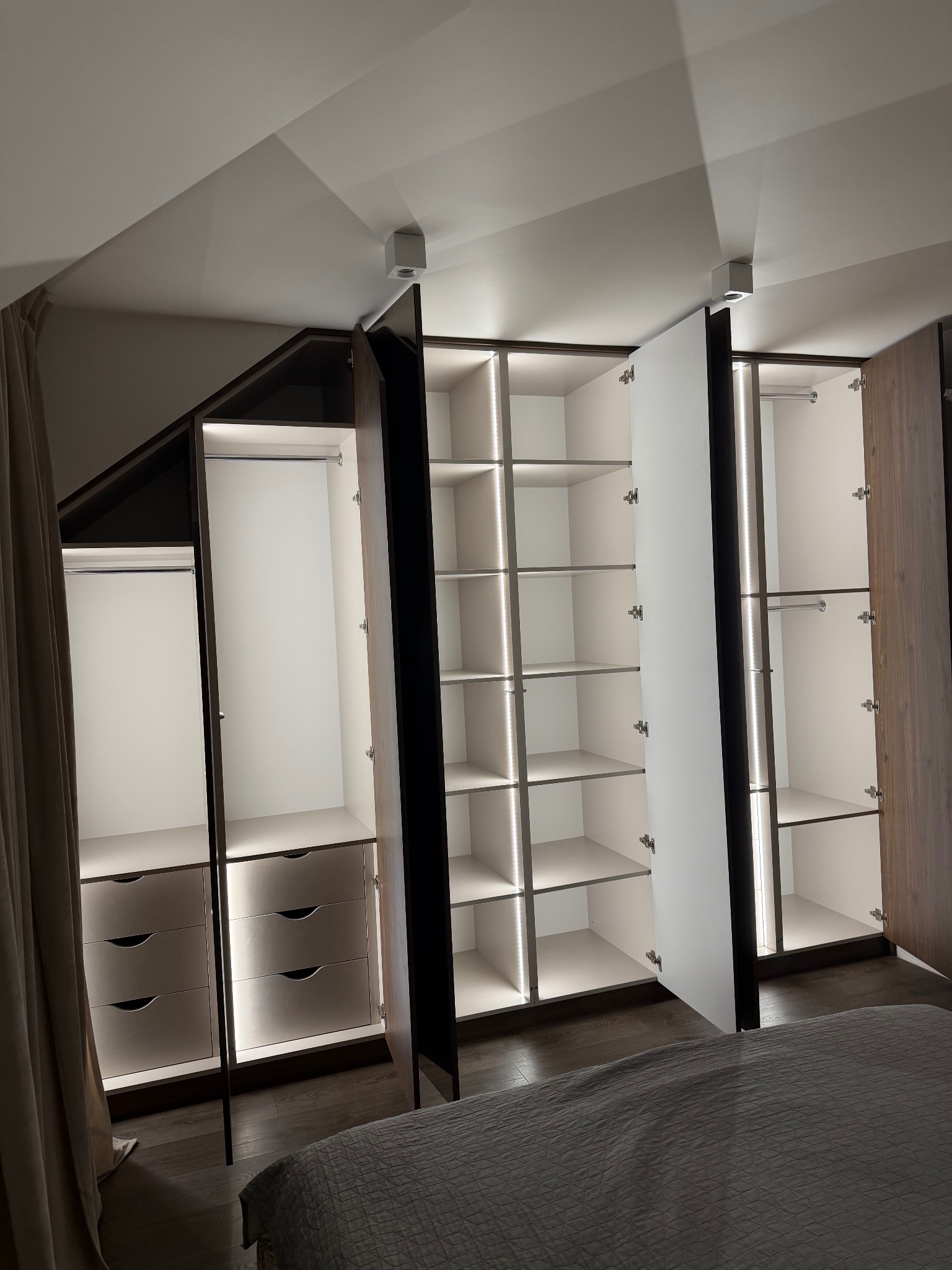 Nowoczesna szafa wnękowa z oświetleniem LED, otwarte drzwi ukazują półki i szuflady. Ciemne drewno kontrastuje z bielą wnętrza. Widoczne detale wykończenia.
