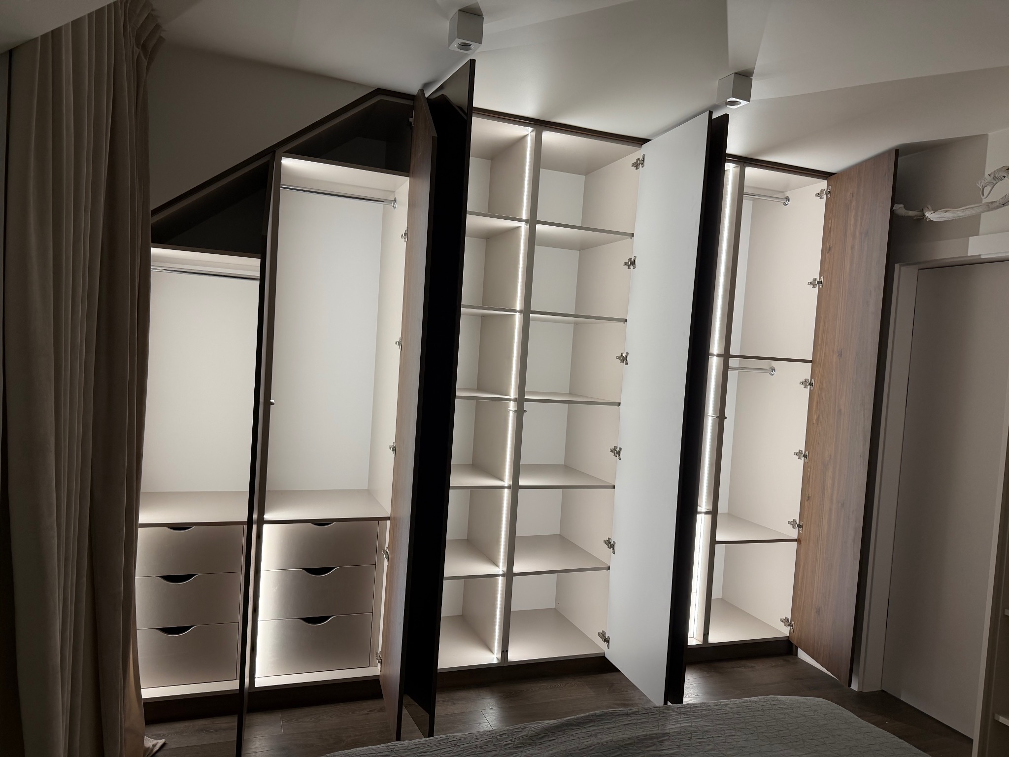 Otwarta szafa wnękowa z oświetleniem LED, widoczne półki i szuflady. Fronty w kolorze białym i drewnopodobnym. Ujęcie z dołu, widoczny fragment łóżka.