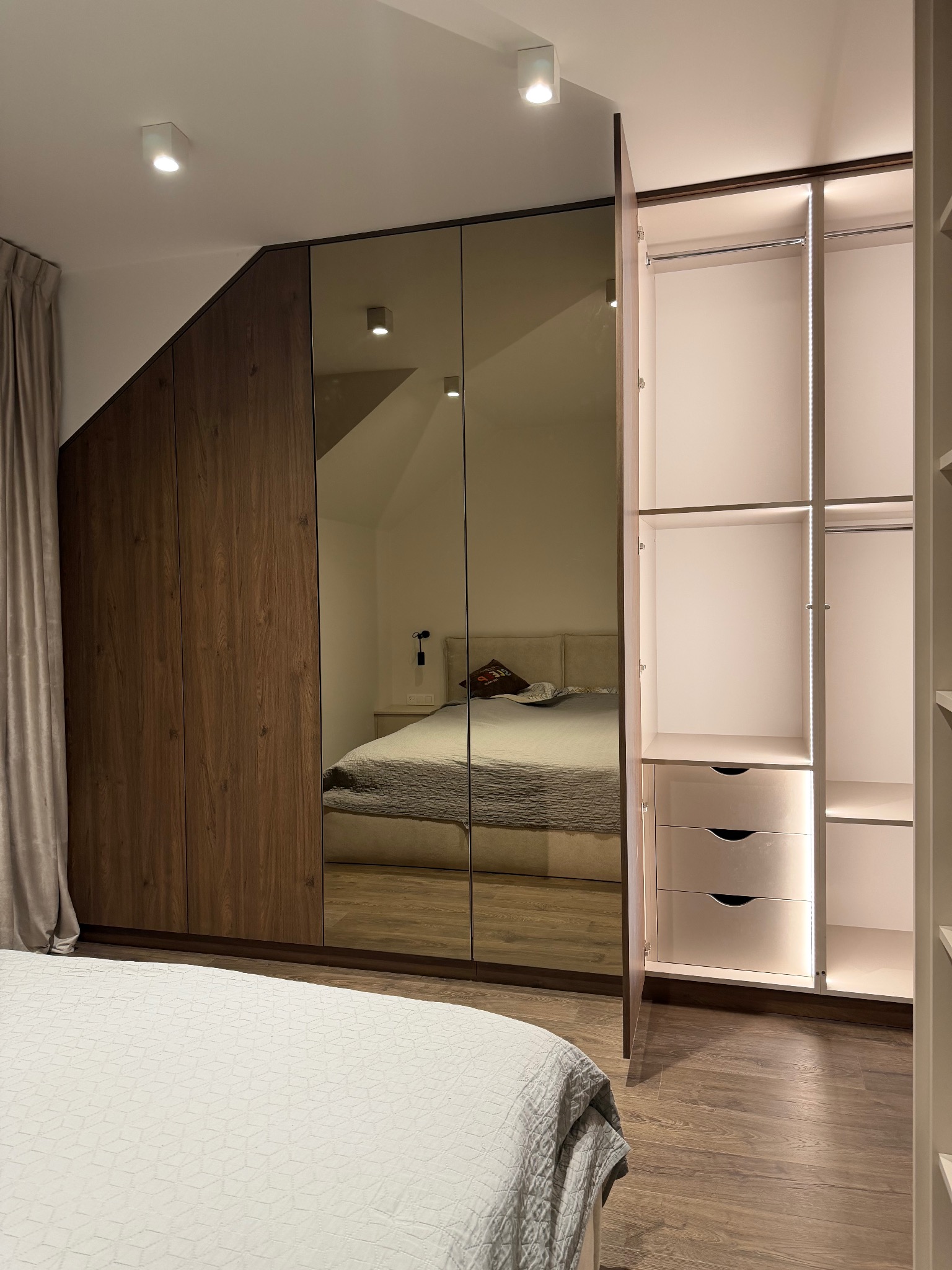 Nowoczesna szafa wnękowa z lustrzanymi drzwiami i oświetleniem LED, dopasowana do skosu dachu. Widoczny fragment sypialni w odbiciu.