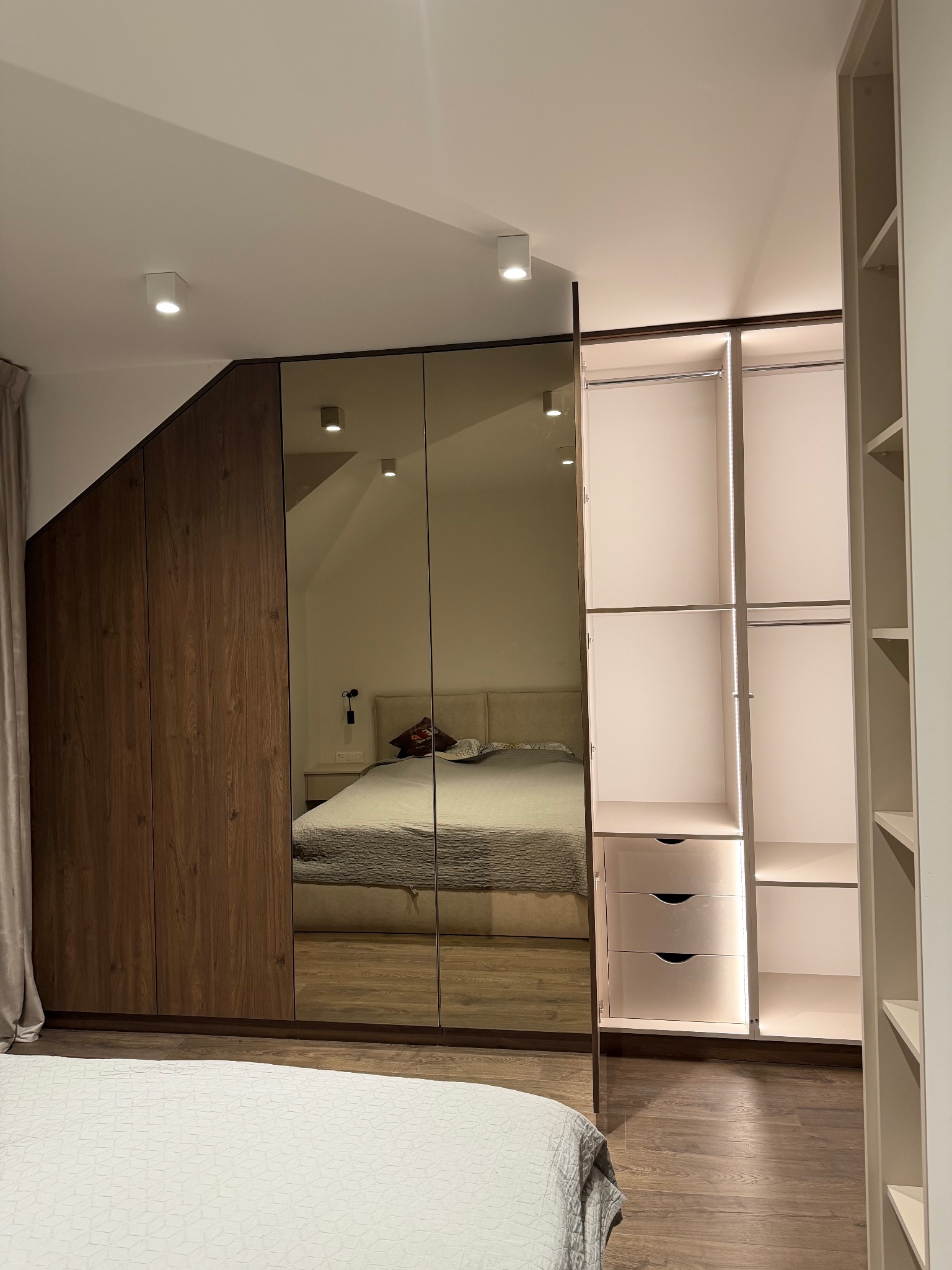 Szafa wnękowa z lustrzanymi drzwiami i drewnianymi bokami, wkomponowana w skos dachu. Oświetlenie LED wewnątrz, widoczna część sypialni w odbiciu.