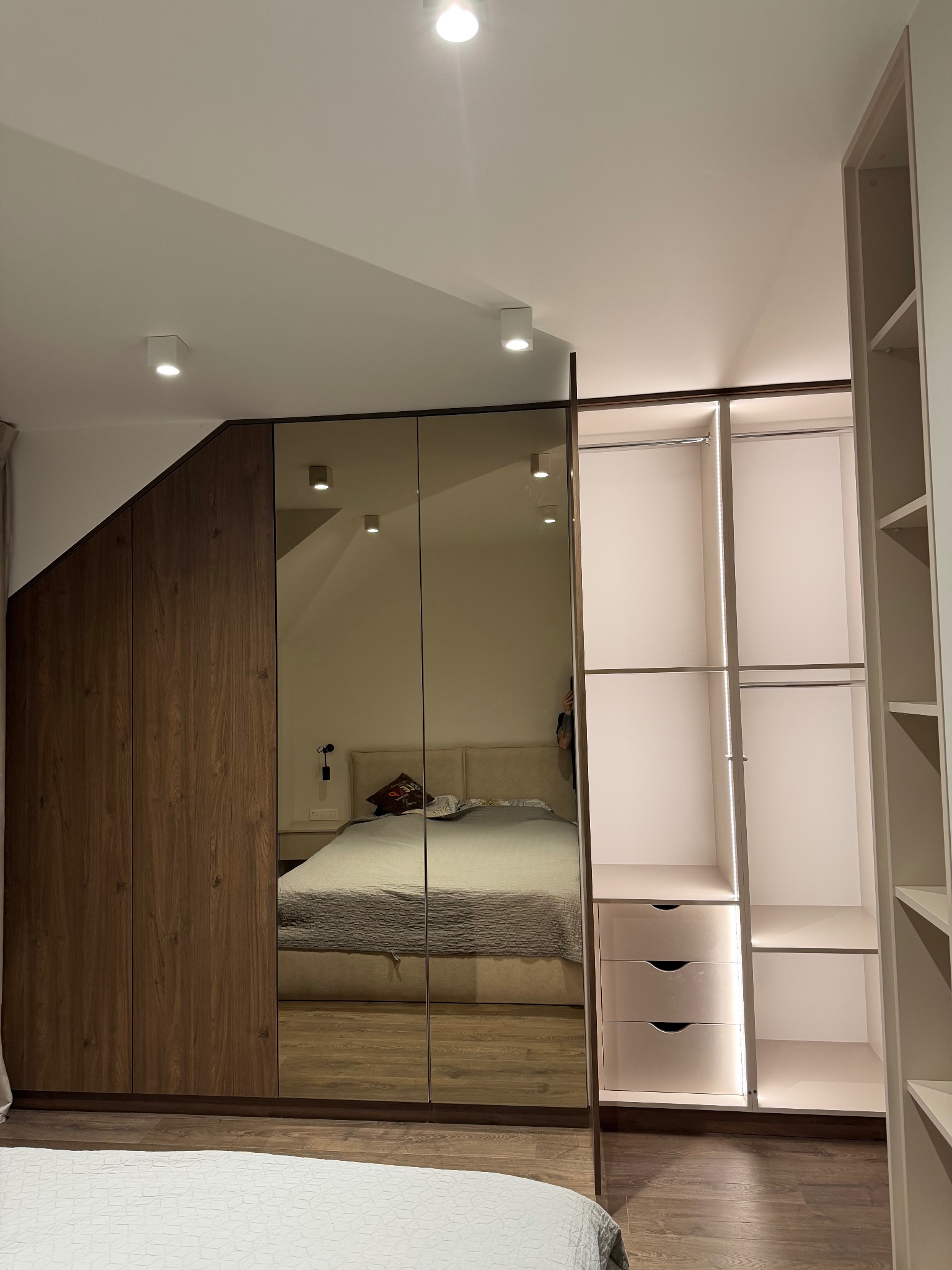 Szafa wnękowa z lustrzanymi drzwiami i oświetleniem LED w sypialni. Widoczne półki i szuflady. Część szafy z drewnianymi frontami pod skosem.