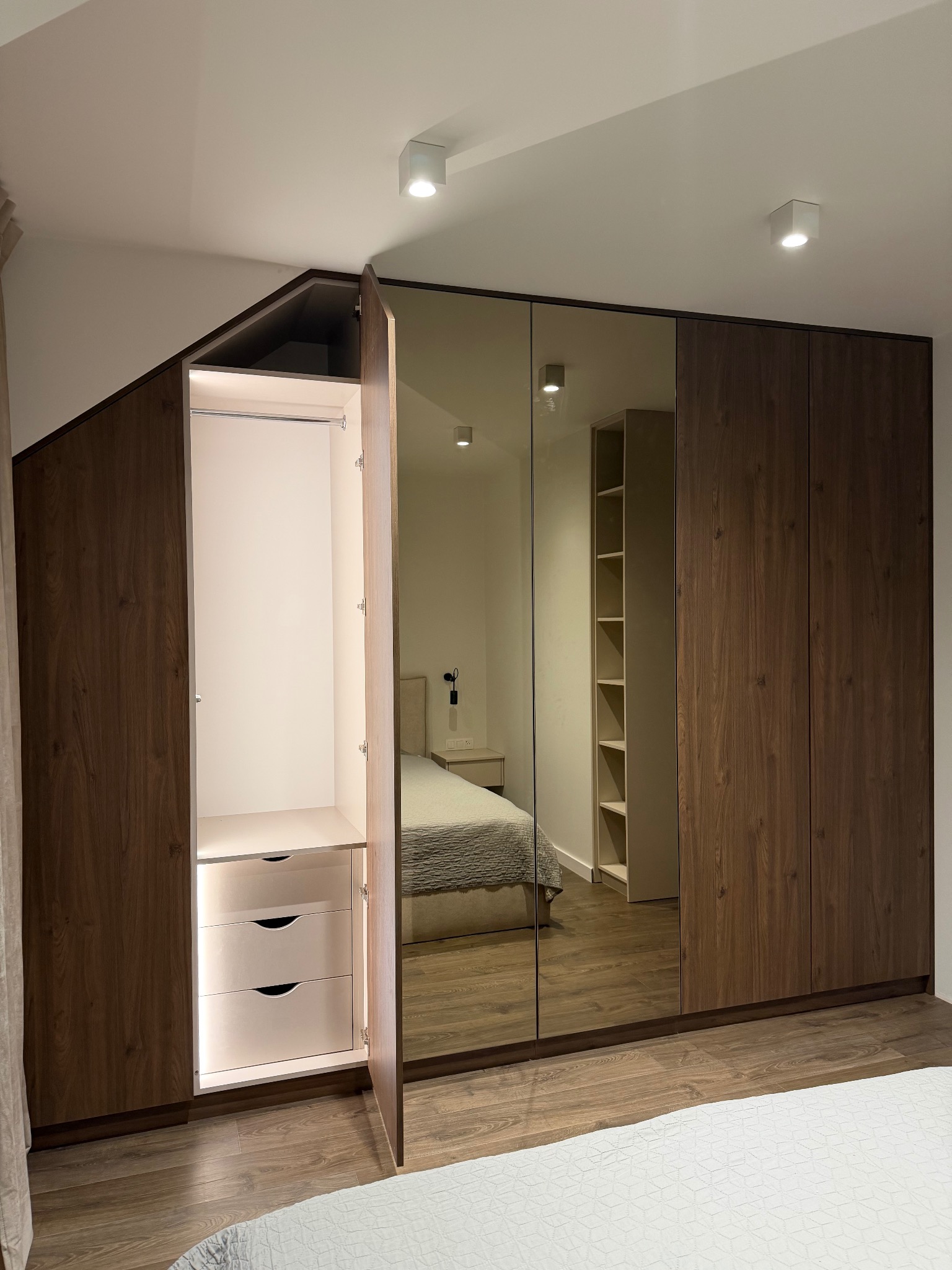 Szafa wnękowa z lustrzanymi drzwiami i oświetleniem LED wewnątrz, z otwartymi drzwiami ukazującymi półki i szuflady. Widoczne odbicie sypialni w lustrze.