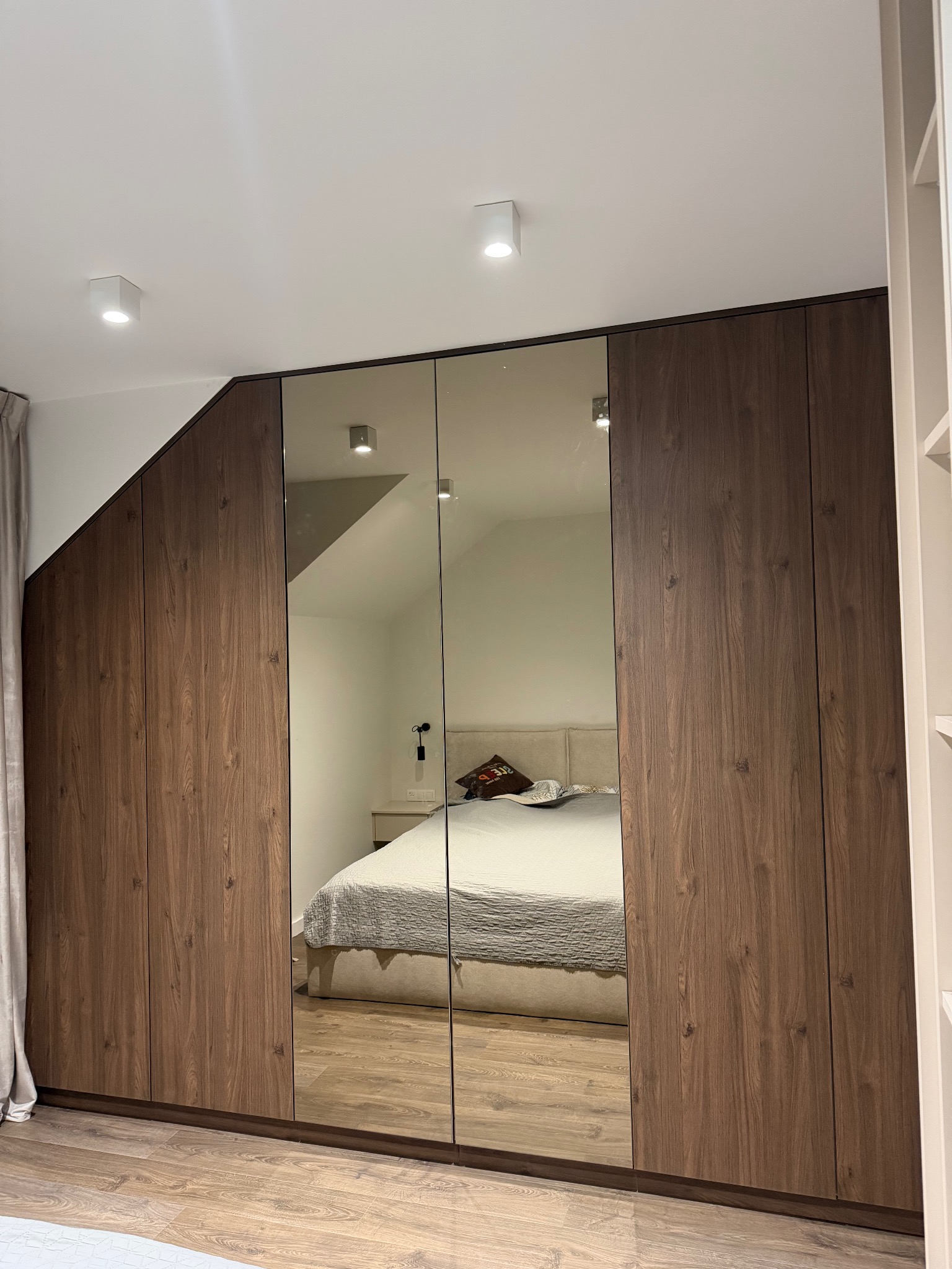 Szafa wnękowa z lustrzanymi drzwiami, drewnopodobne fronty w odcieniach brązu, dopasowana do skosu dachu, odbicie sypialni w lustrach.