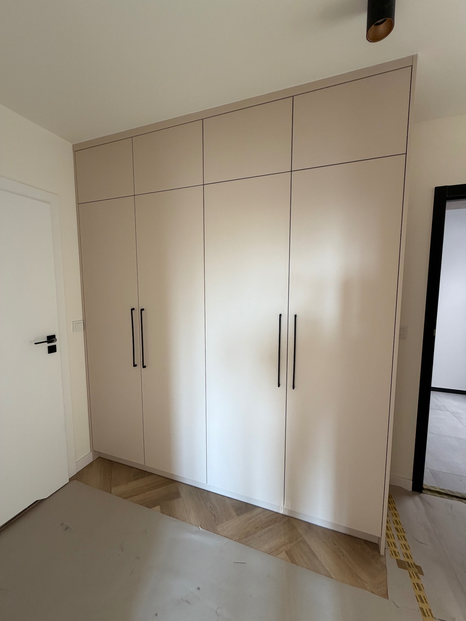 Beżowa szafa wnękowa z czarnymi uchwytami, dopasowana do wnętrza z parkietem w jodełkę. Minimalistyczny design, widok na fragment drzwi i sufit z lampą.