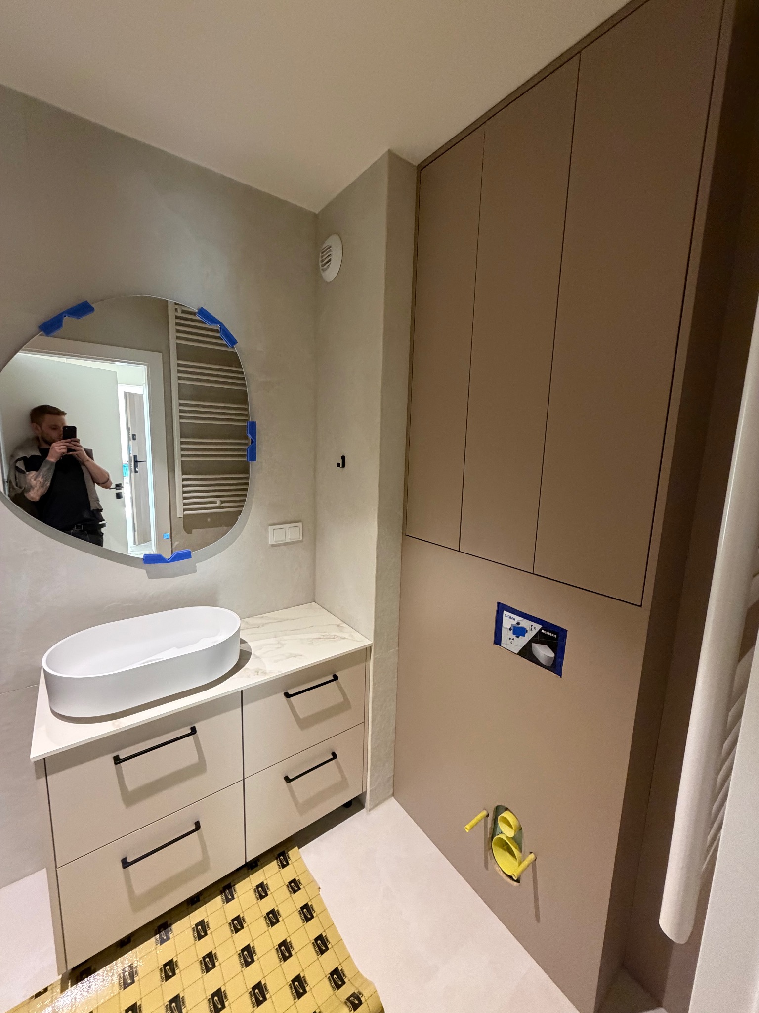 Nowoczesna łazienka w beżach: owalne lustro z podświetleniem, umywalka nablatowa na szafce z szufladami i wysoka zabudowa. Widoczne elementy instalacji.