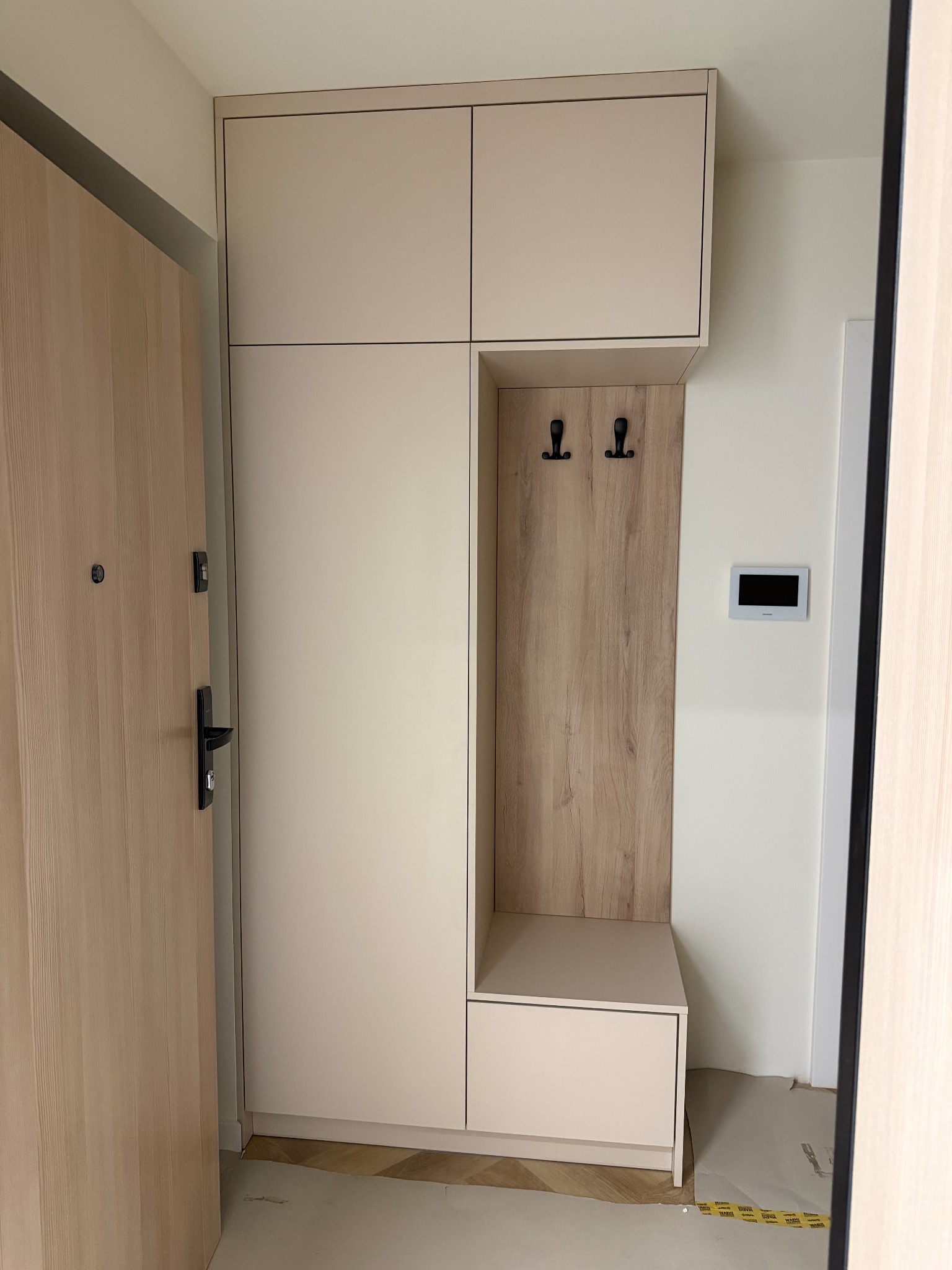 Jasna szafa na wymiar z wnęką na wieszaki i siedziskiem, dopasowana do przedpokoju. Minimalistyczny design, drewnopodobne akcenty, widoczna część drzwi wejściowych.