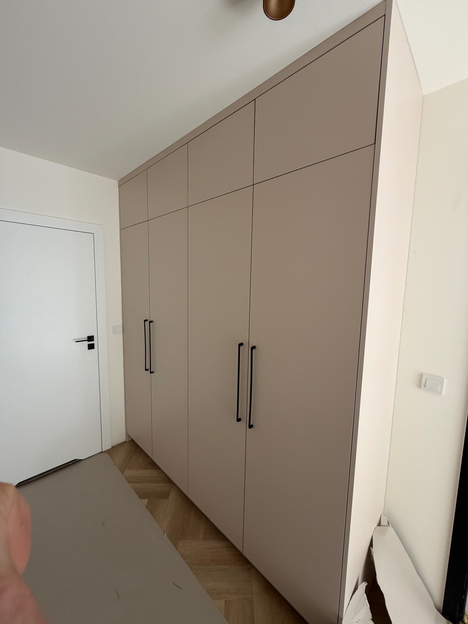 Beżowa szafa wnękowa z czarnymi, długimi uchwytami, dopasowana do jasnego wnętrza z parkietem. Minimalistyczny design, widoczne detale wykończenia.