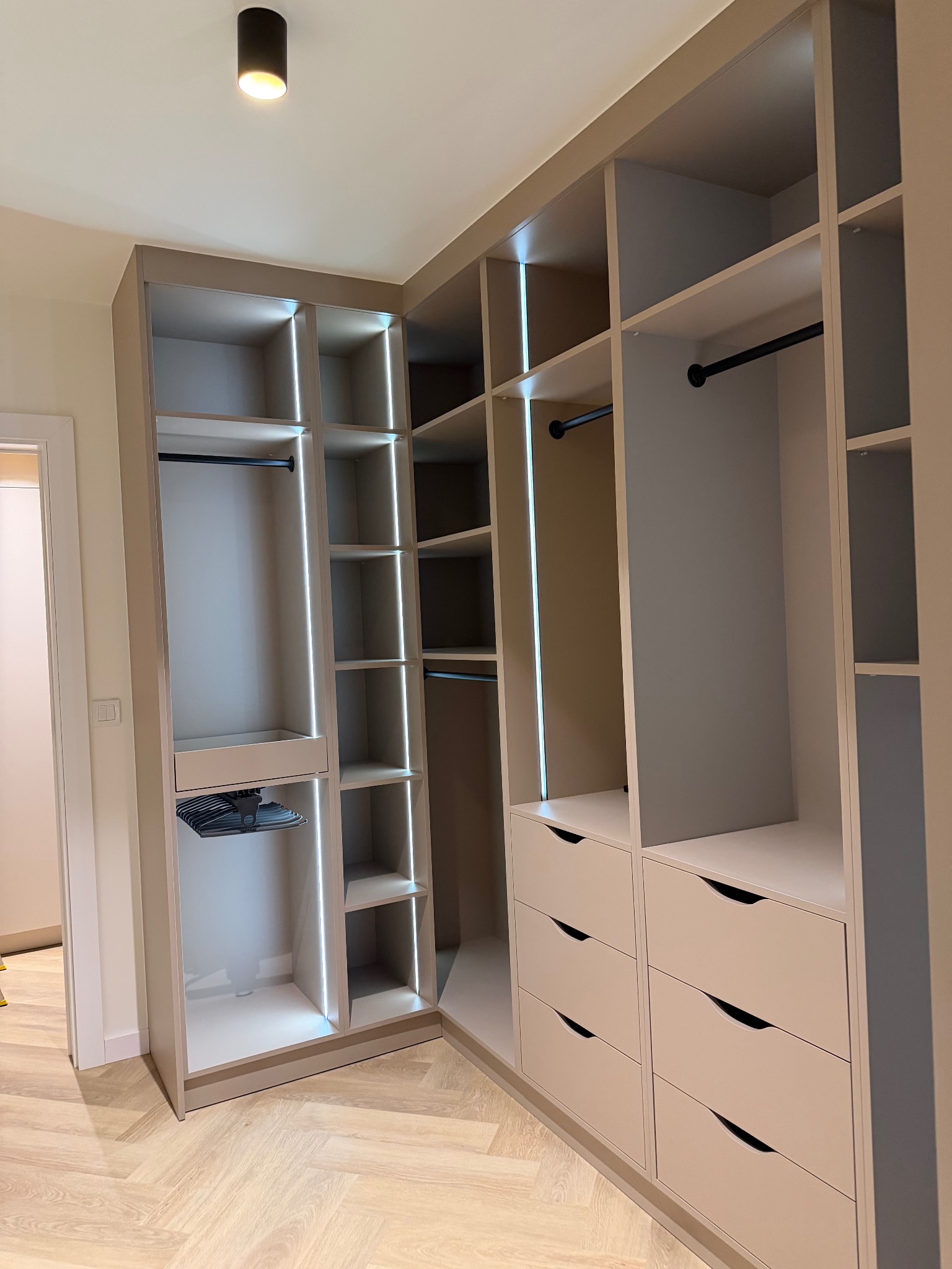 Jasna, narożna szafa wnękowa z oświetleniem LED, widoczne półki, drążki i szuflady z charakterystycznymi uchwytami. Podłoga z parkietu w jodełkę.