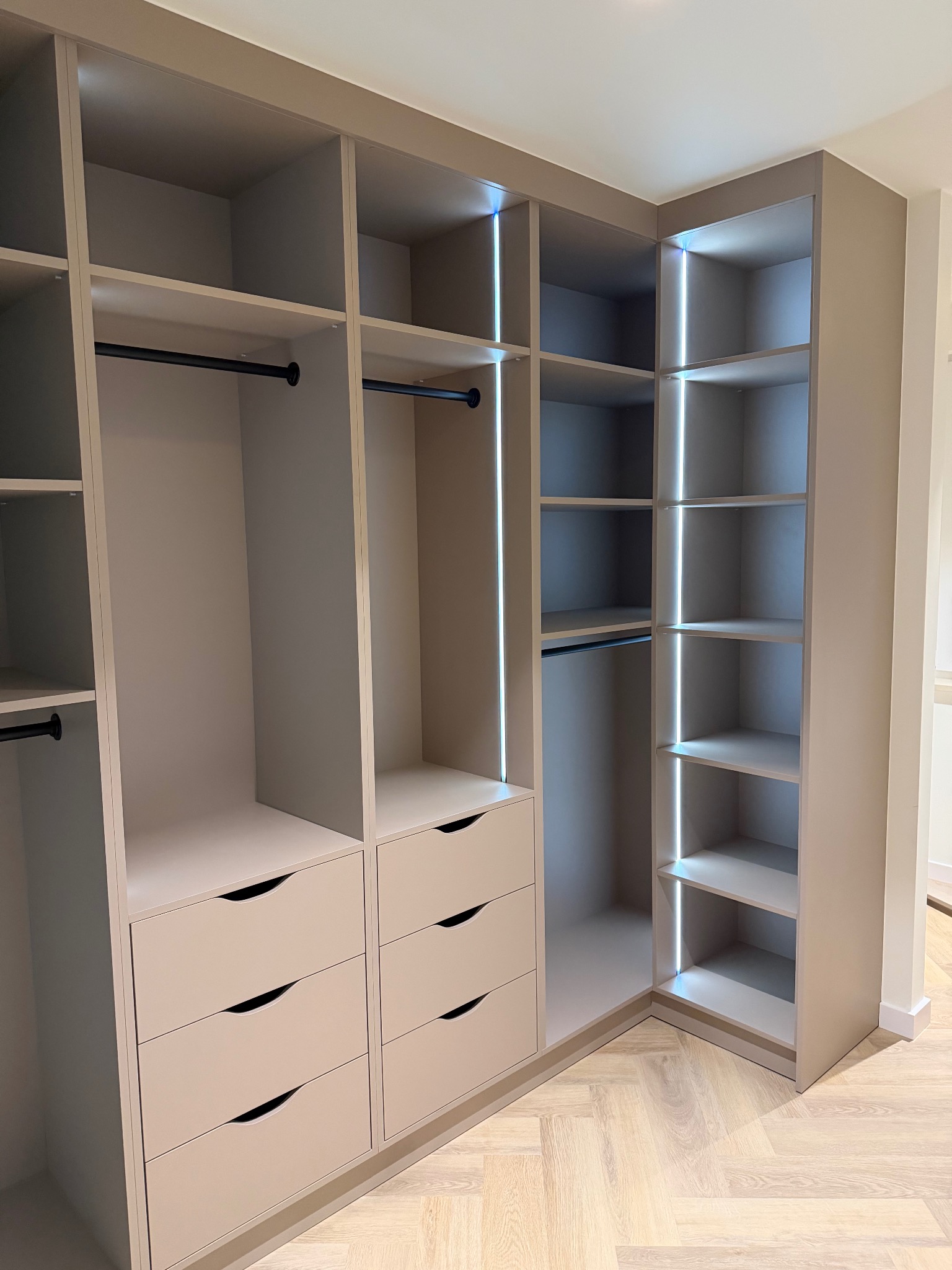 Jasna, narożna szafa wnękowa z szufladami i oświetleniem LED, widok z bliska na półki i drążki na ubrania. Parkiet w jodełkę na podłodze.