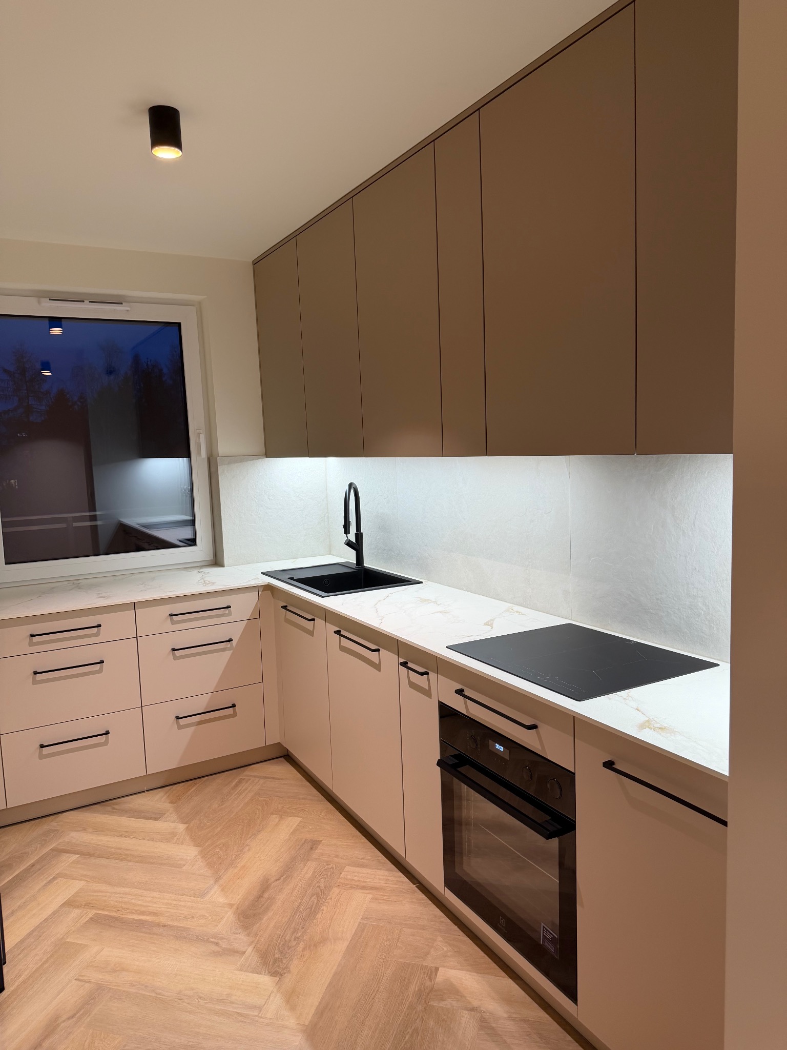 Nowoczesna kuchnia z beżowymi frontami, czarnymi uchwytami i armaturą. Blat imitujący marmur, podświetlenie LED i parkiet w jodełkę tworzą elegancki wystrój.