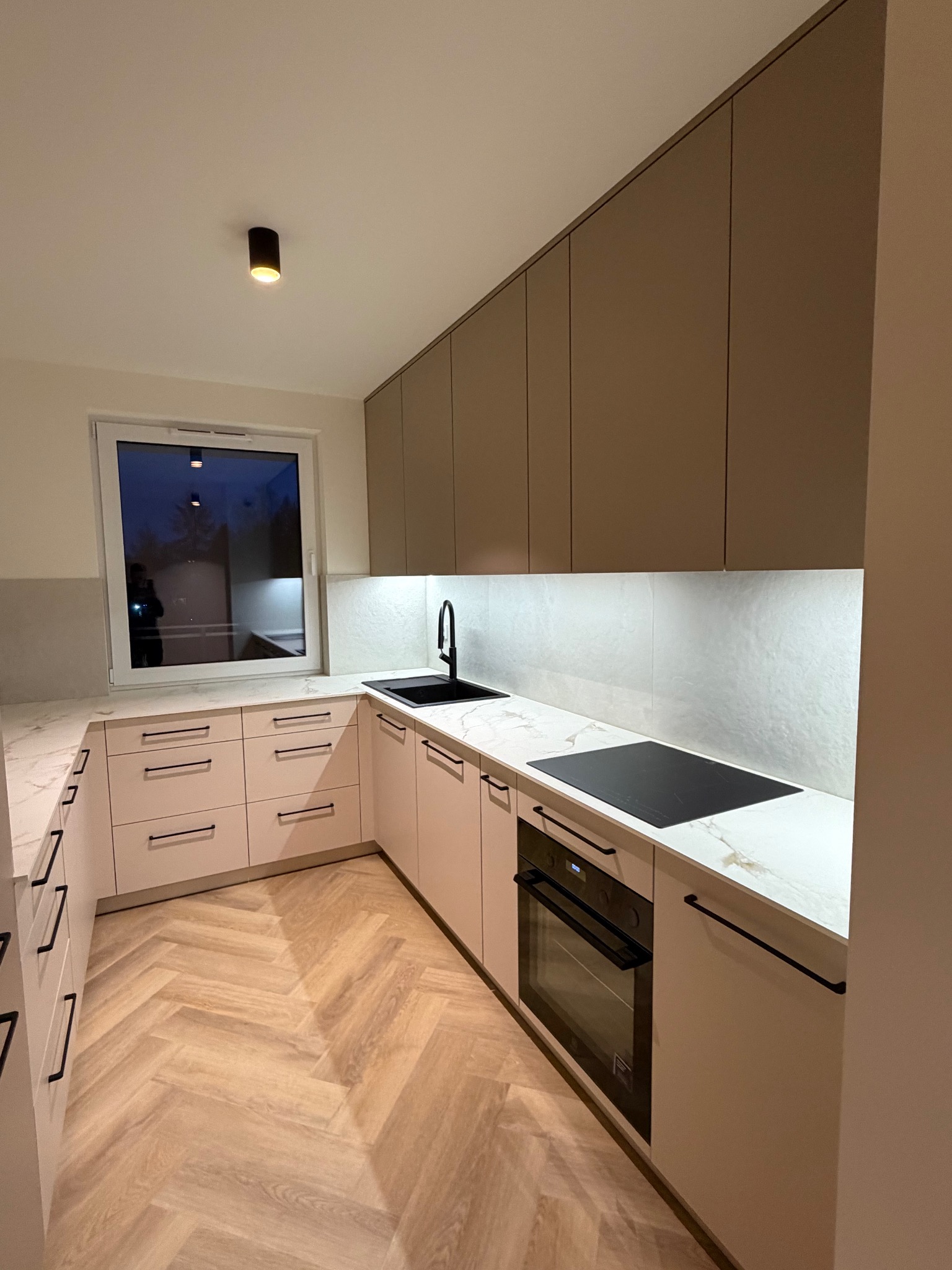 Nowoczesna kuchnia w kształcie litery U z beżowymi frontami, czarnymi uchwytami i blatem imitującym marmur. Na blacie czarna płyta indukcyjna i zlew. Parkiet ułożony w jodełkę.