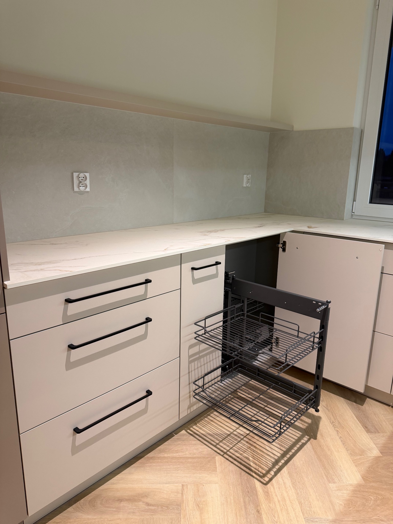 Jasna kuchnia z wysuwanym koszem cargo. Fronty szafek w odcieniach beżu, czarne uchwyty. Na ścianie gładka, beżowa płytka. Parkiet ułożony w jodełkę.