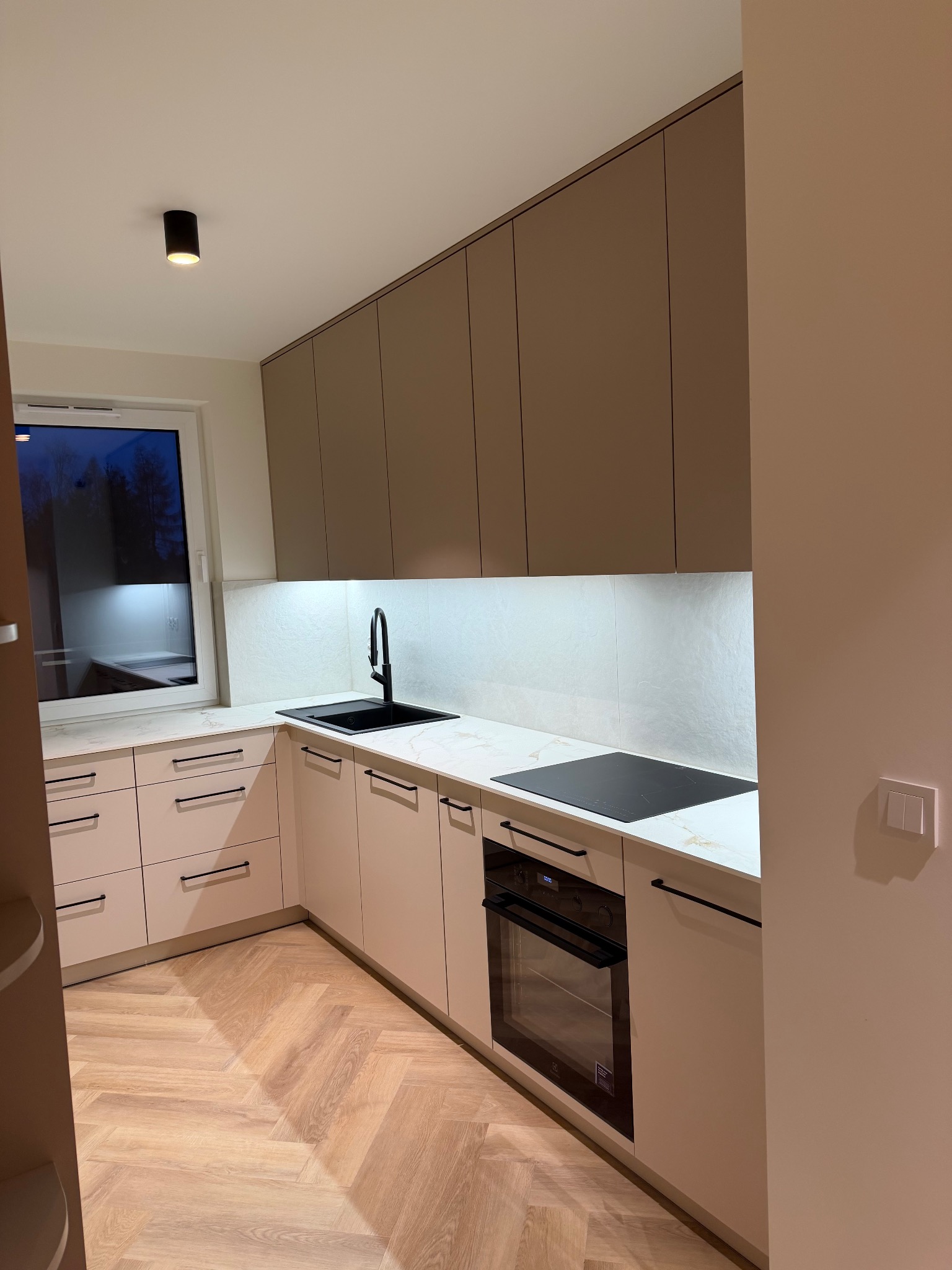 Nowoczesna kuchnia na wymiar z beżowymi frontami, czarną armaturą i blatem imitującym marmur. Widoczna płyta indukcyjna i piekarnik. Parkiet ułożony w jodełkę.