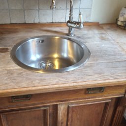 Vitalii Furman - Rustykalny blat drewniany z wbudowanym zlewozmywakiem i baterią. Widoczne szafki pod blatem, dekoracje i płytki na ścianie w tle.