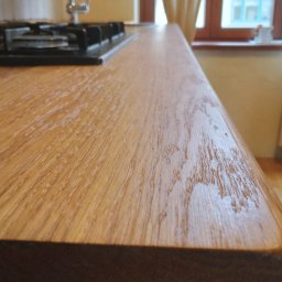 Vitalii Furman - Drewniany blat kuchenny z zaokrąglonym brzegiem, widoczny fragment płyty grzewczej i okno w tle. Naturalne oświetlenie podkreśla strukturę drewna.